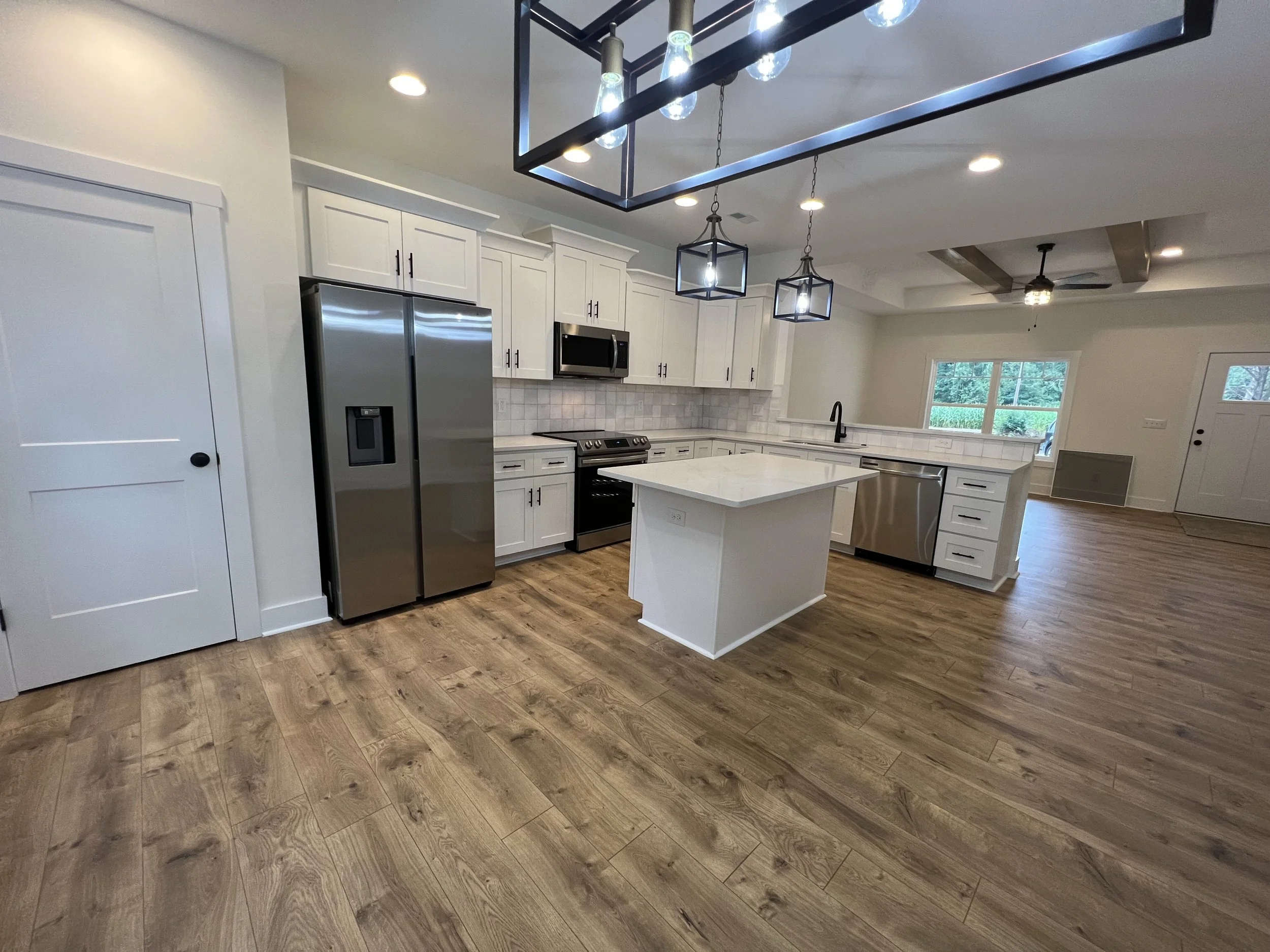 Modern kitchen with white cabinets, stainless steel refrigerator and oven, quartz island, and hanging black geometric pendant lights, LVP hardwood floors, large window with outdoor view.