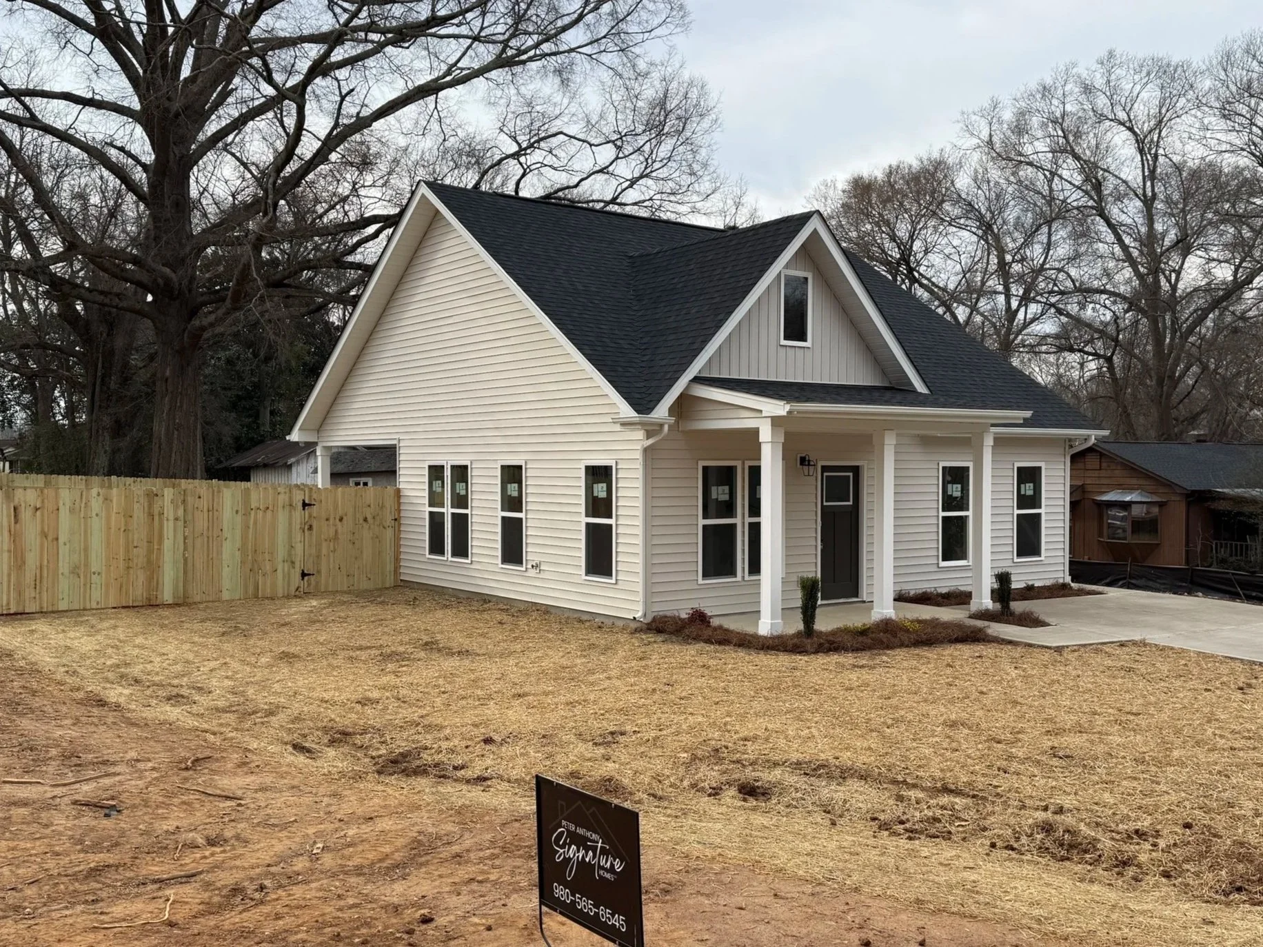 New construction herringbone and white trim house with Certainteed black shingle roof, large covered front porch and double hung windows, fenced backyard, and surrounded by trees.