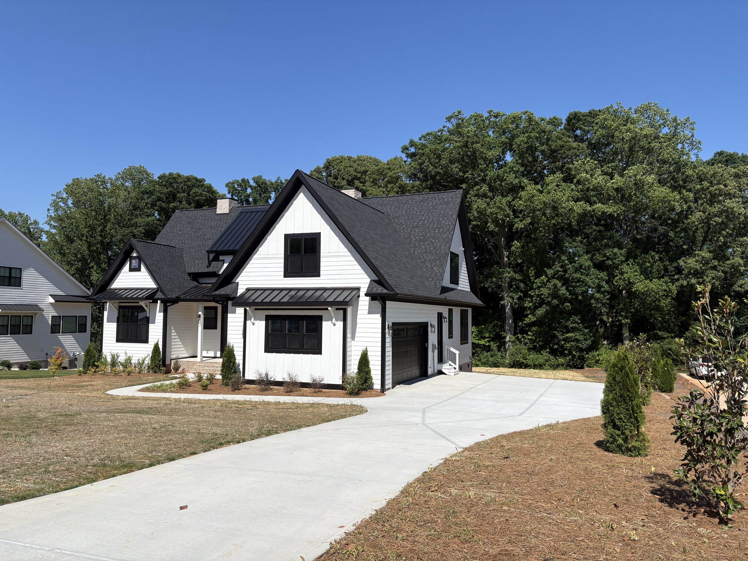 A modern white house with black trim and a black roof, surrounded by a concrete driveway and patches of grass and shrubs in the yard. Tall green trees are in the background under a clear blue sky.