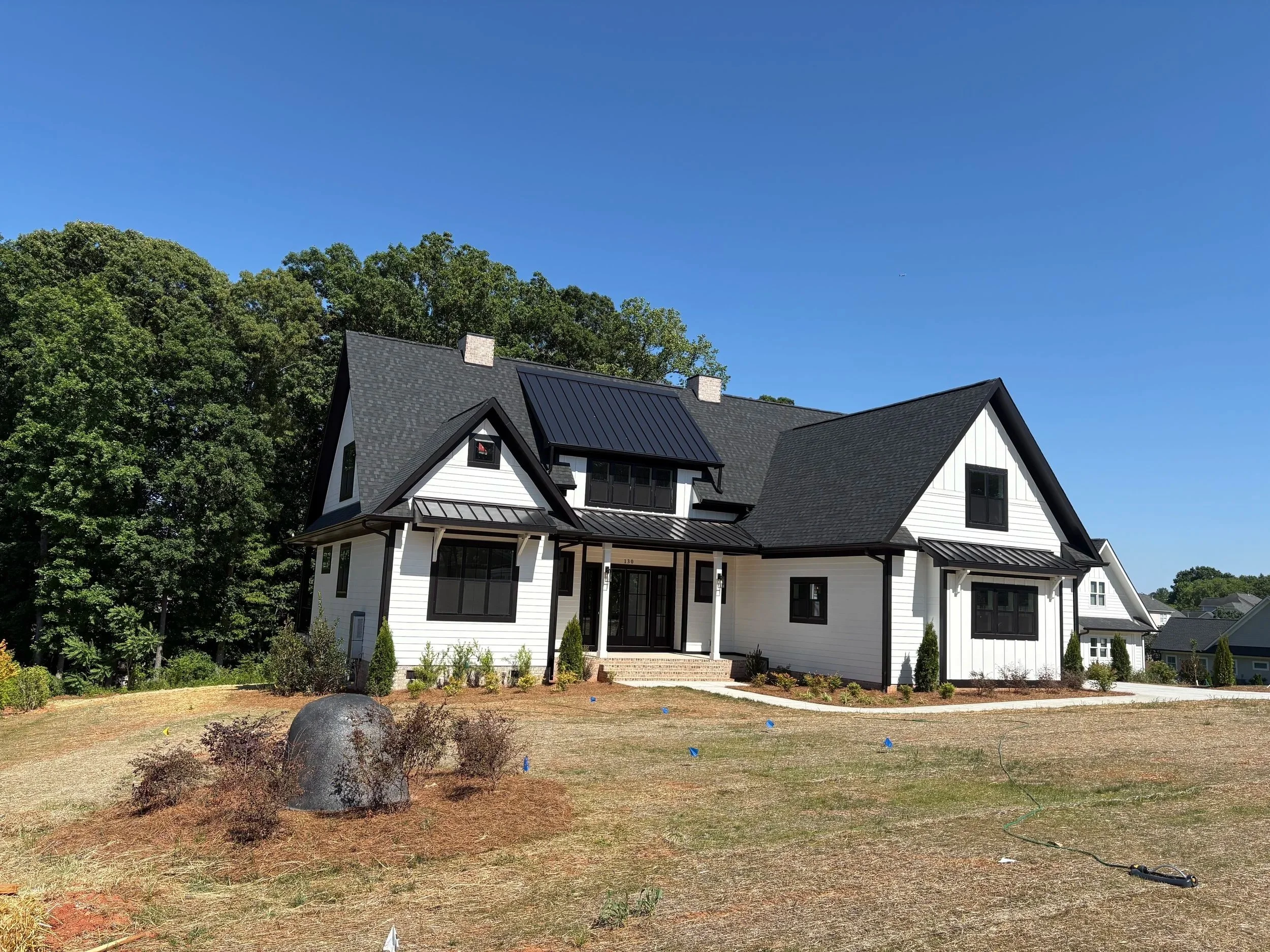 A modern two-story farmhouse with white siding, black window frames, and a black Certainteed roof, surrounded by trees and plants and shrubs.