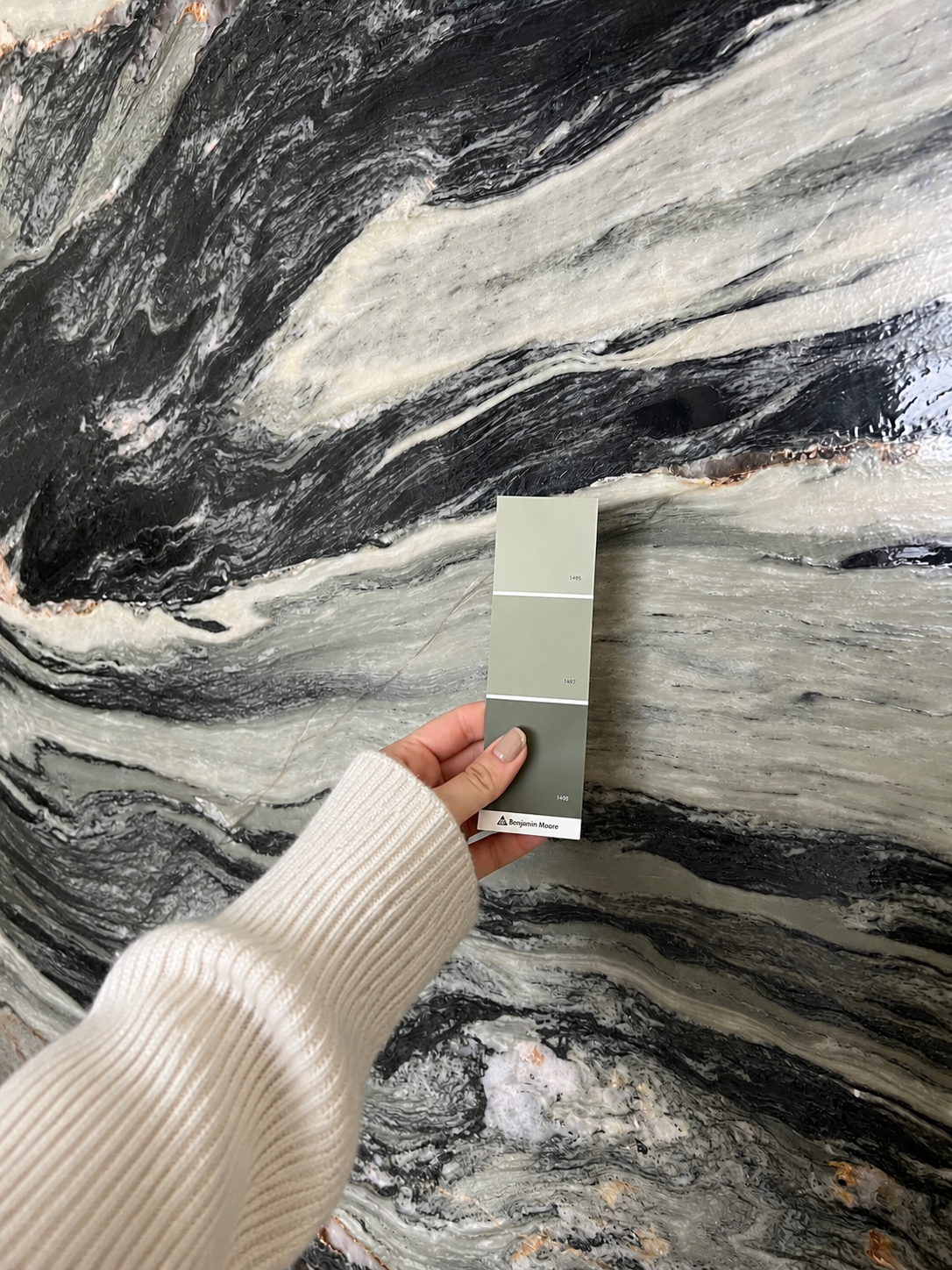 Person holding a color sample card in front of a large patterned marble or stone wall with black, white, and gray swirling veins.