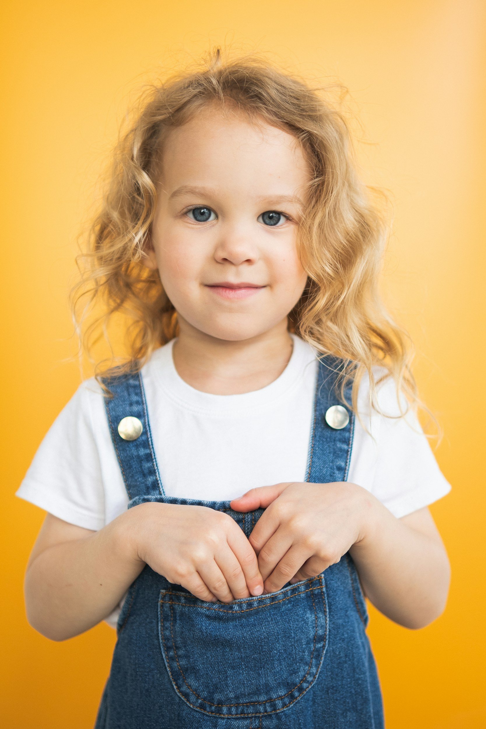 Young girl with curly blonde hair and blue eyes, wearing a white t-shirt and denim overalls, standing against a yellow background, with her hands clasped together