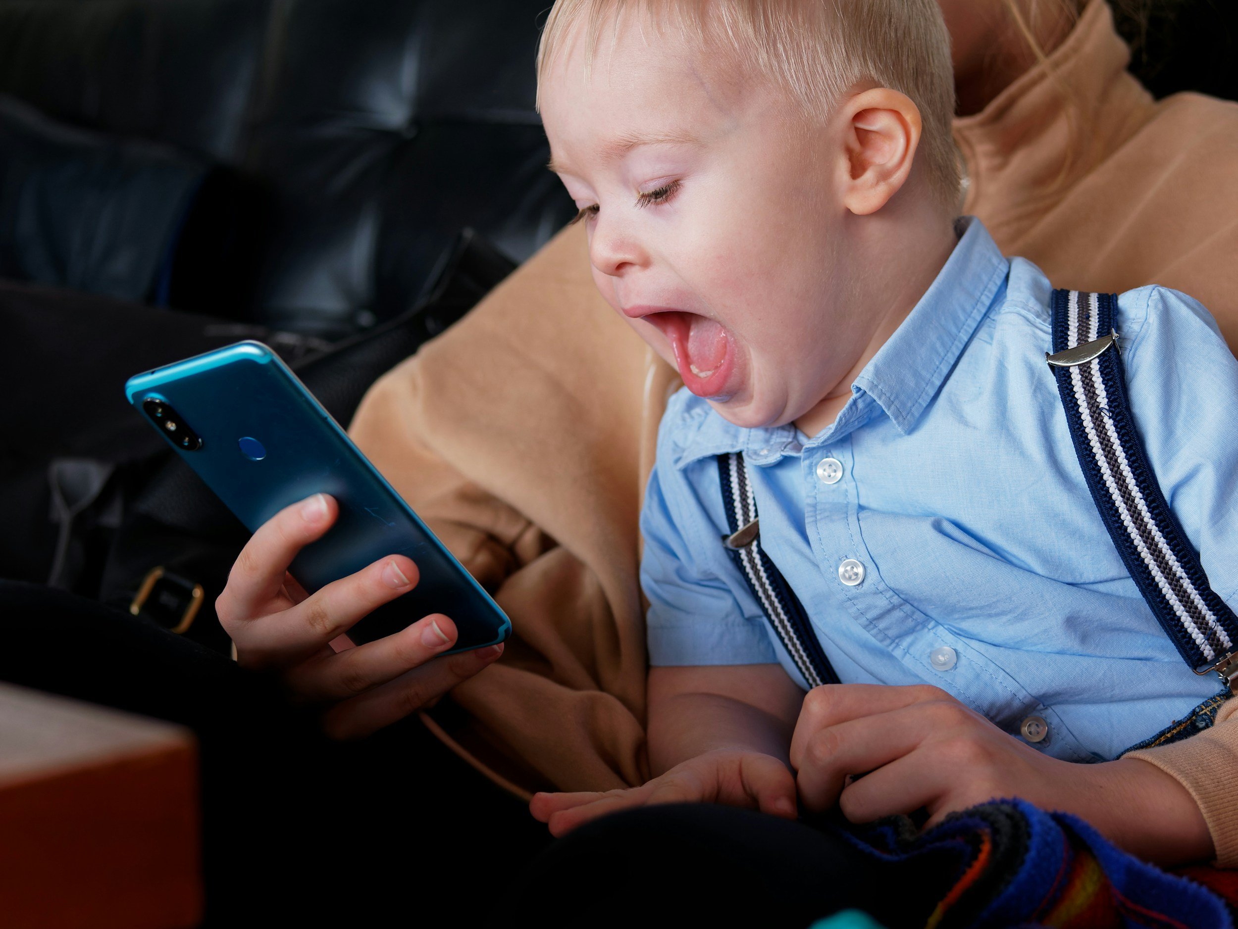 A young blond boy in a blue shirt playing on a smartphone, lying on a person's lap while yawning or shouting.