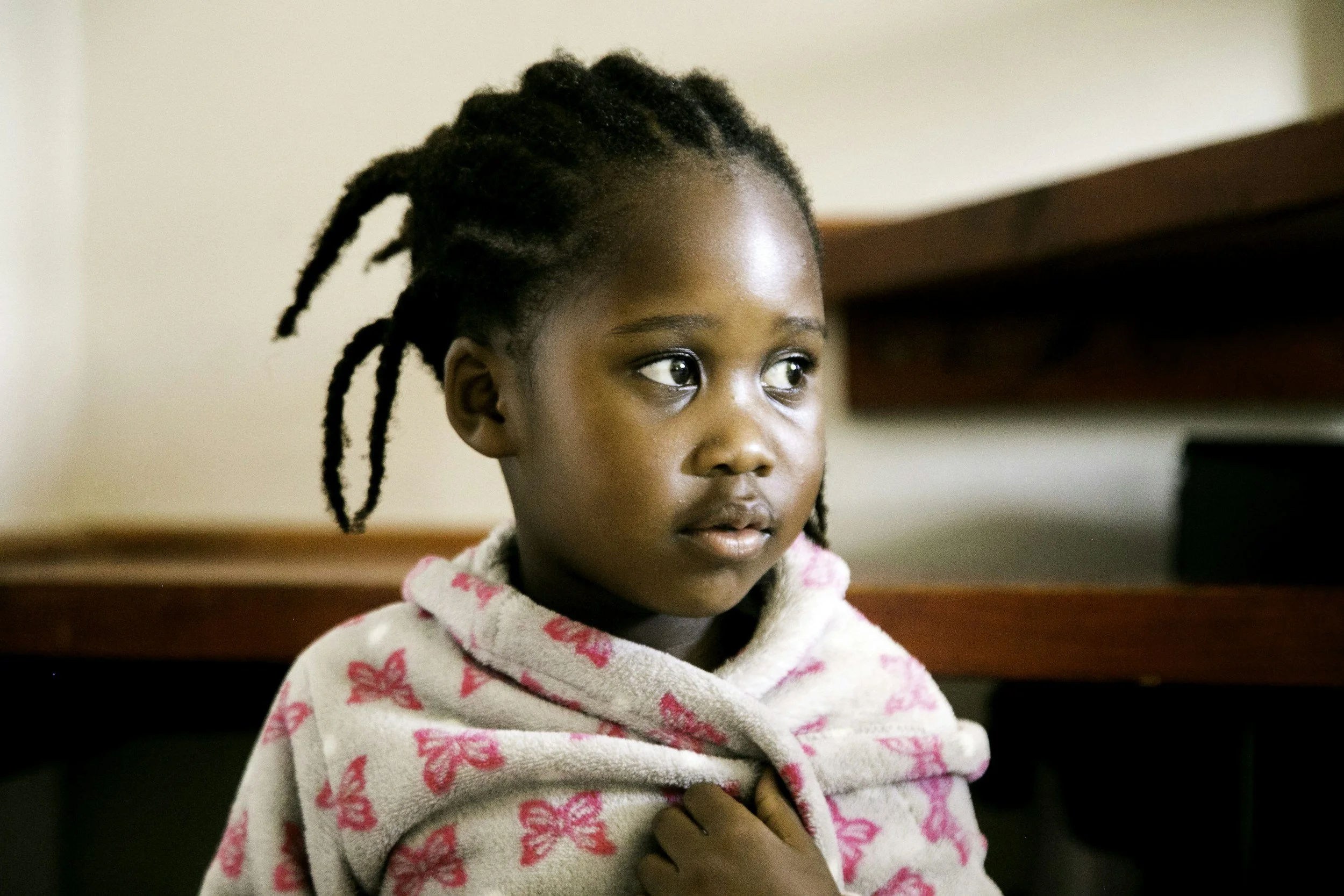 Close-up of a young girl with dreadlocks looking to the side indoors, wearing a hooded sweatshirt with pink butterfly patterns, standing near a wooden surface.