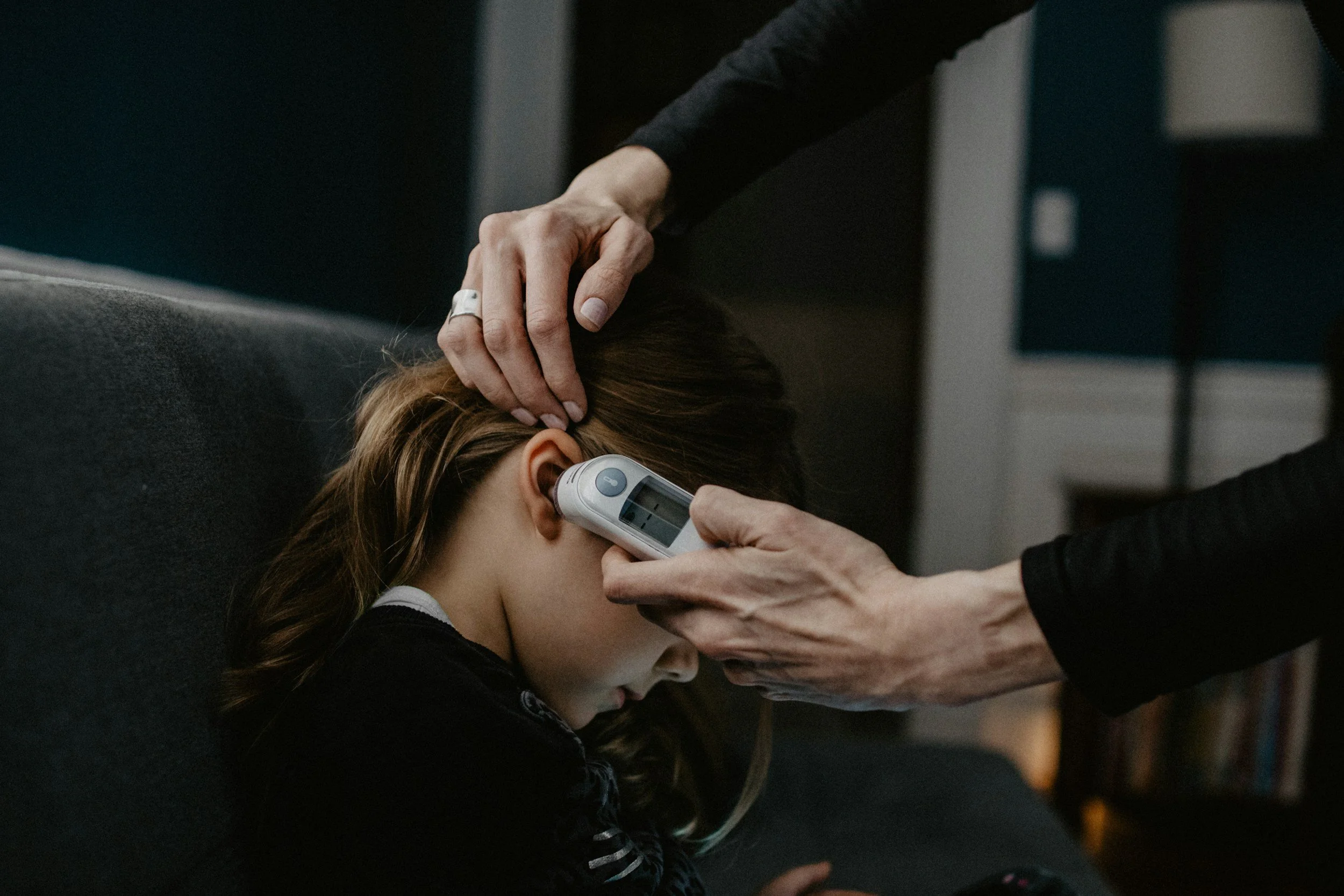 A young girl with long hair resting her head against a surface while a person checks her temperature with a thermometer, with a hand gently placed on her head.