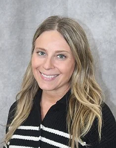 Portrait of a smiling woman with long blond hair wearing a black and white sweater against a gray background.