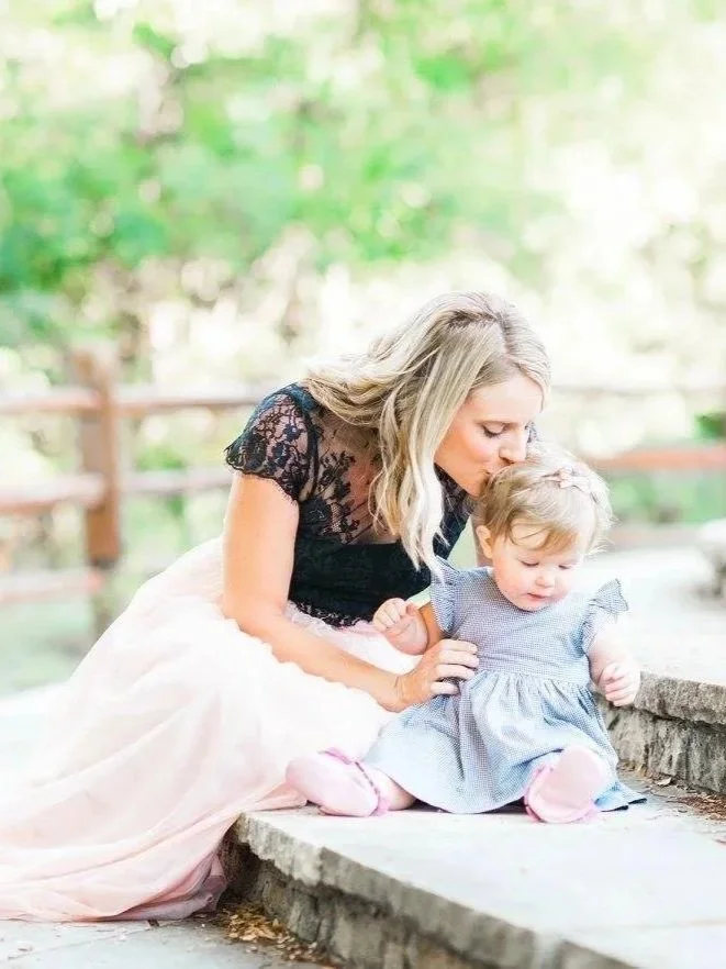 A woman with blonde hair in a black lace top and pink skirt kneels on the ground, kissing a young girl with blonde hair dressed in a blue dress, sitting on a stone ledge in a park with green trees in the background.