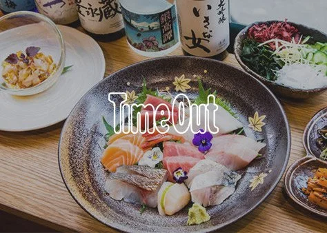 Sashimi platter with various slices of raw fish, garnished with flowers and wasabi, surrounded by small bowls of dipping sauces and side dishes, with bottles of sake in the background.