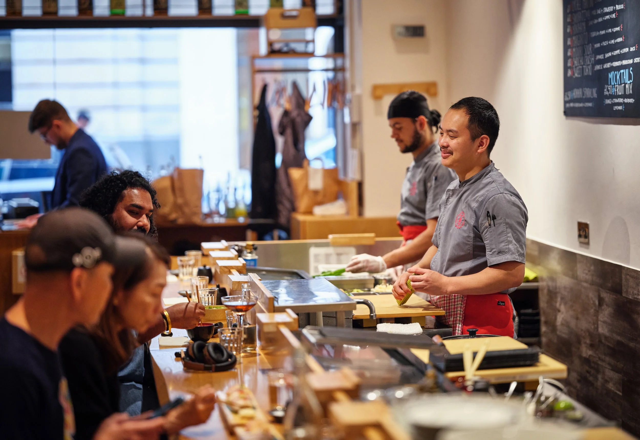 Sushi chefs preparing food behind a counter in a busy restaurant with customers seated in front.
