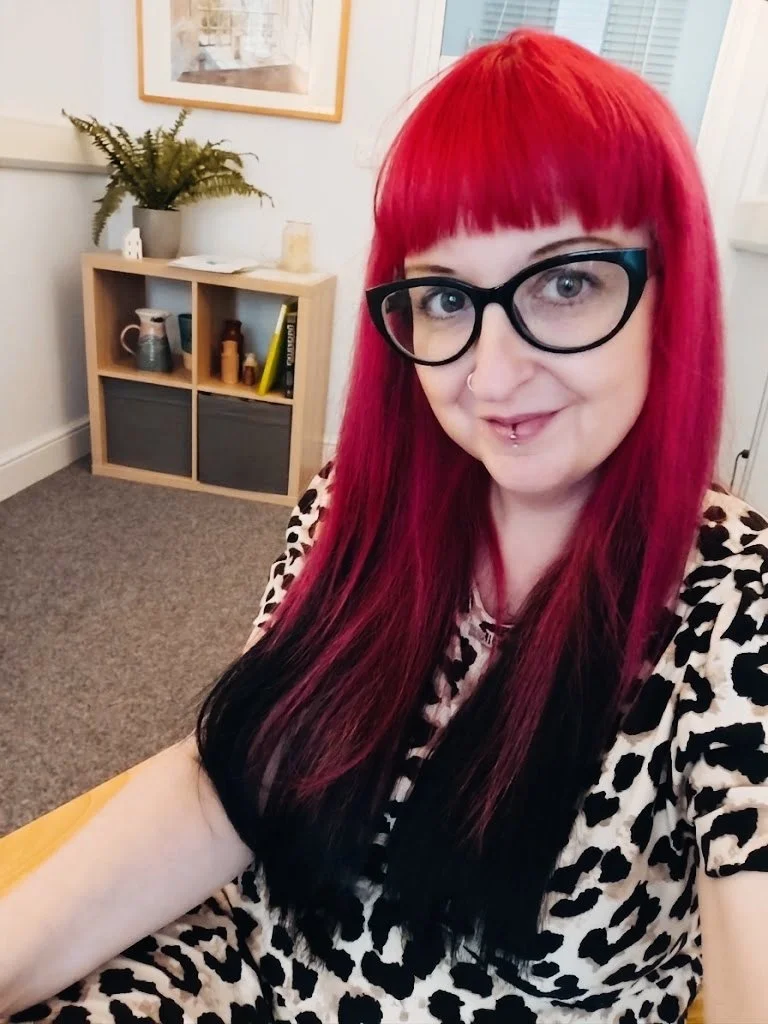 Woman with bright red and black hair, wearing glasses and a leopard print top, sitting in a room with a bookshelf, potted fern, and framed artwork on the wall.