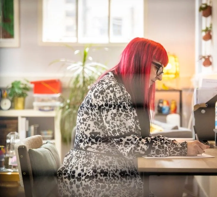 A woman with red hair and glasses working at a desk in a well-lit office.