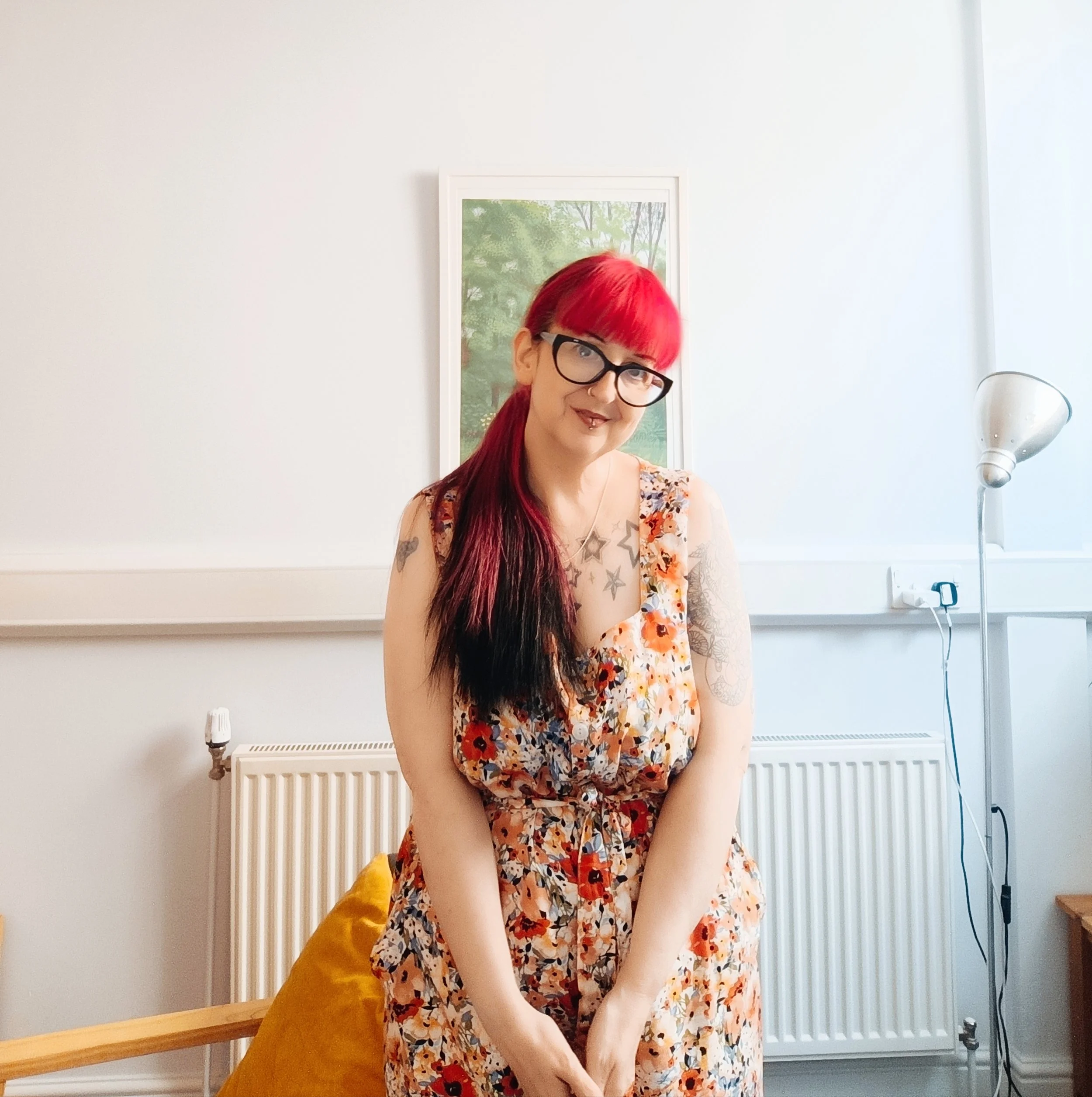 Tara is in this photo. They have bright red hair, glasses, and tattoos, are wearing a floral jumpsuit and sitting on a yellow chair in a room with white walls, a picture of a green forest, a radiator, and a floor lamp.
