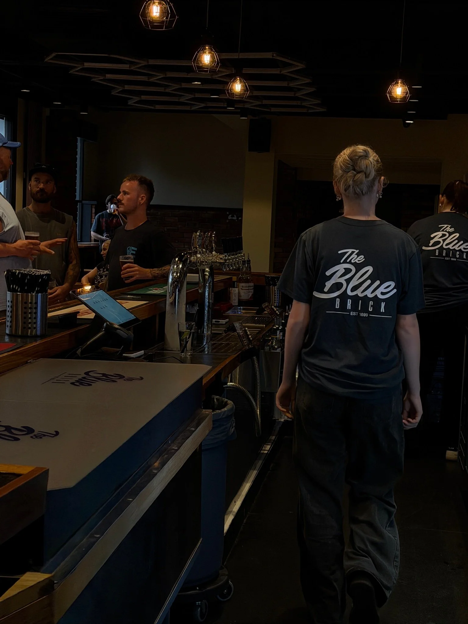 People inside a bar with dark lighting, some talking and holding drinks, and a woman with blonde hair styled in an updo wearing a dark t-shirt that says 'The Blue Brick'.