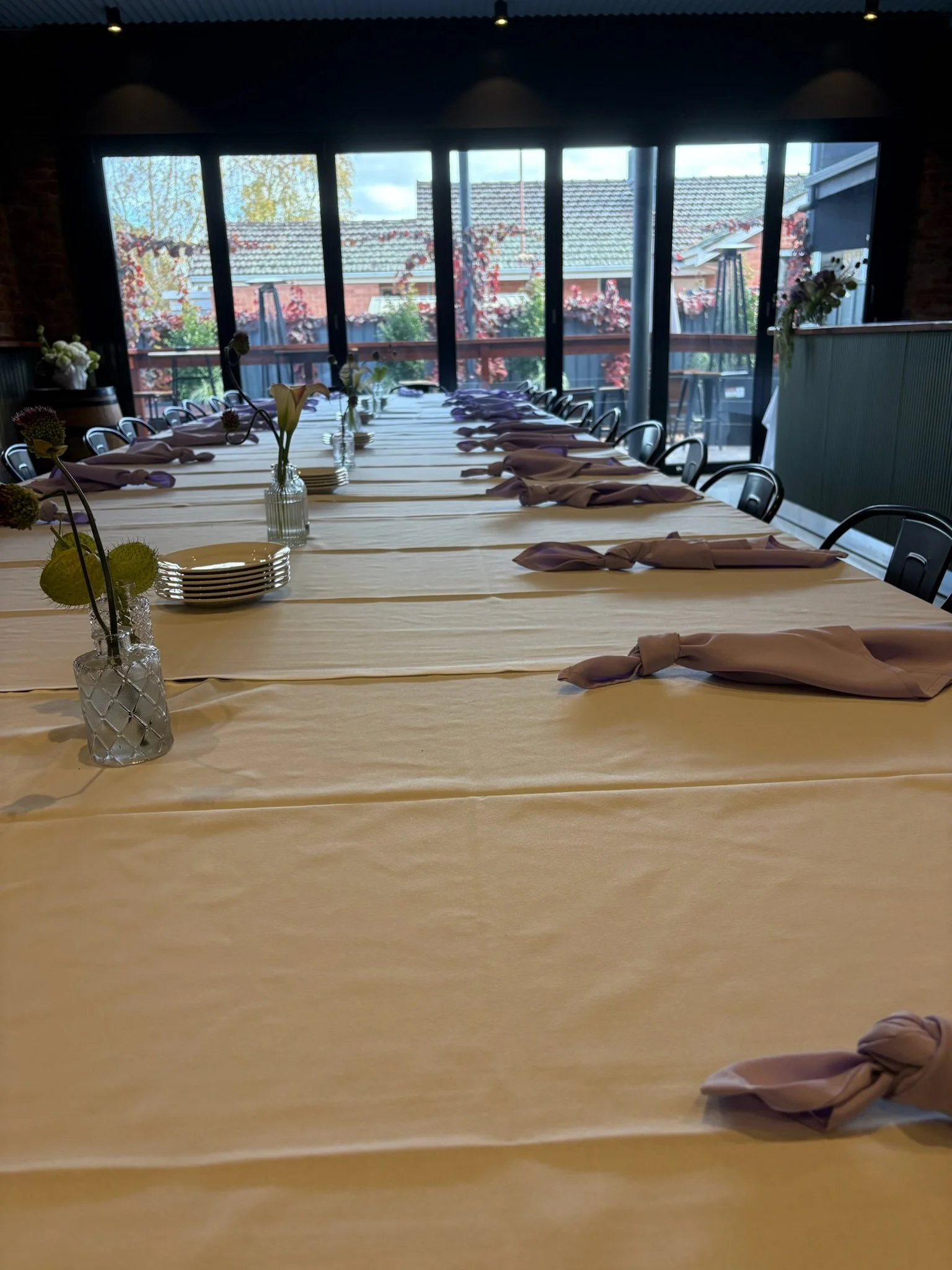 Long banquet table covered with a beige tablecloth, set with plates, napkins, and floral centerpieces in vases, in a room with large windows showing a backyard with trees and rooftops.
