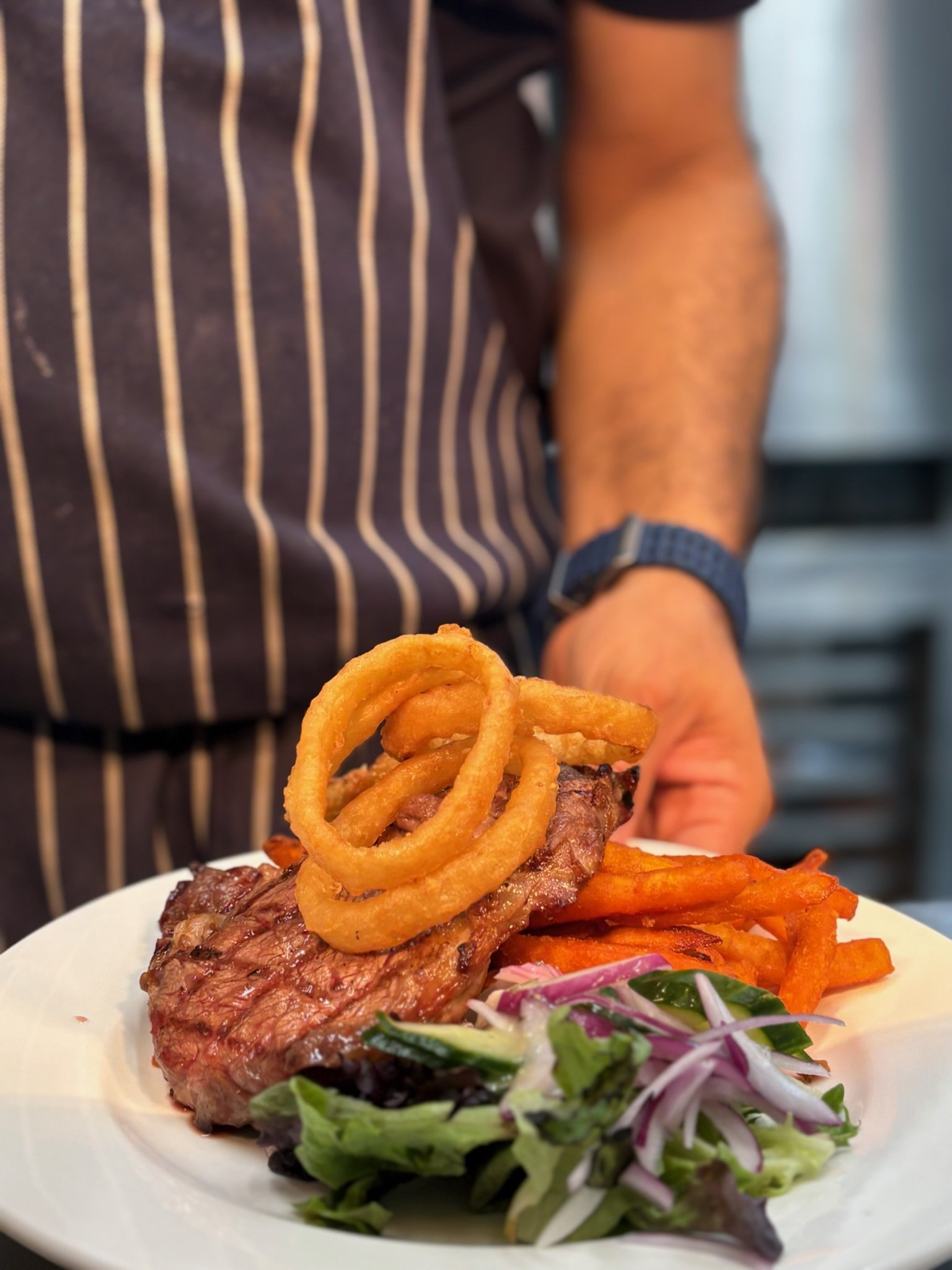 Close-up of a plate with grilled steak topped with onion rings, side salad with greens and sliced vegetables, and crispy sweet potato fries. A person wearing a striped apron and wristwatch is serving the food in the background.