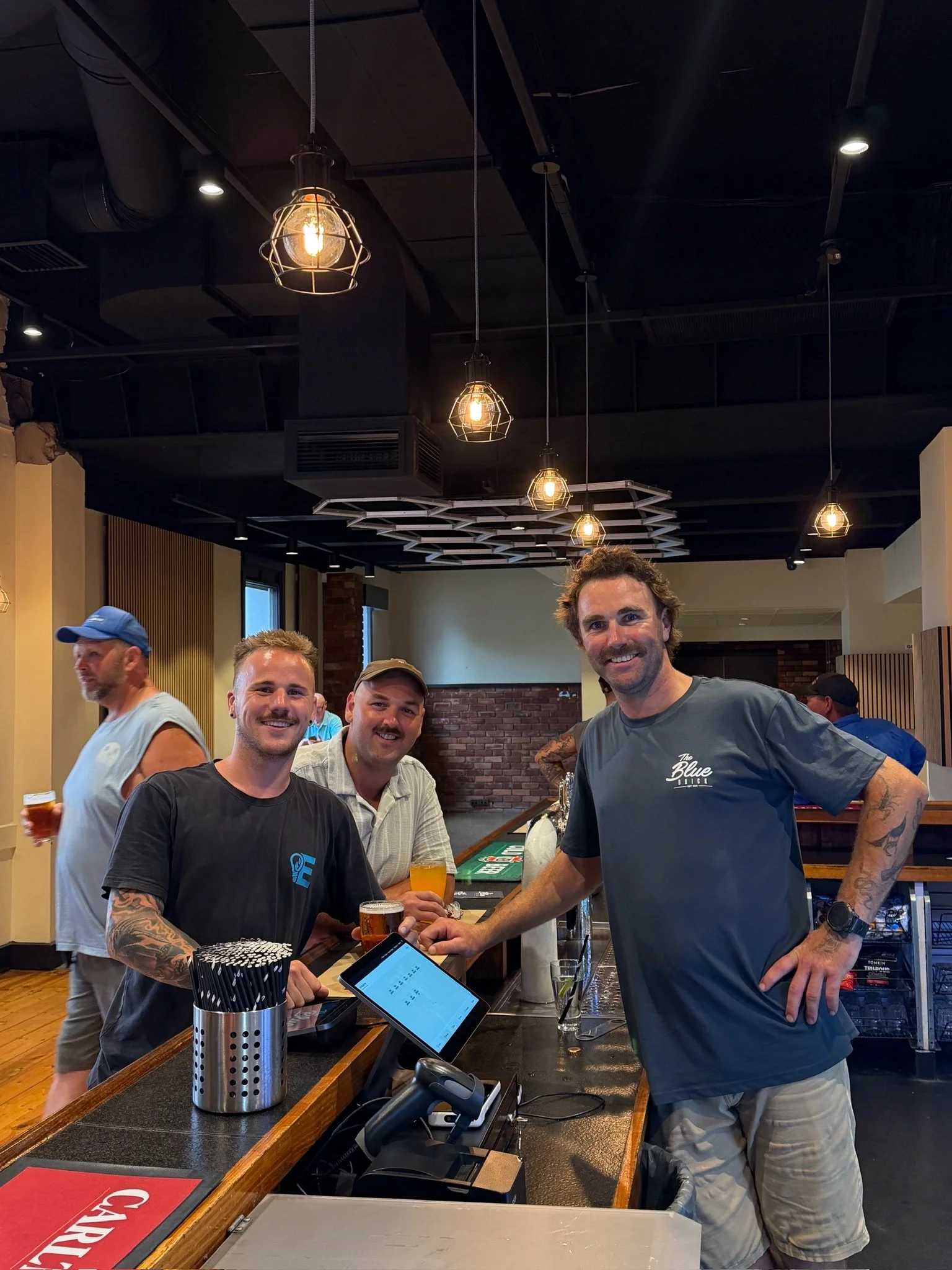 Four men smiling at a bar, with one man wearing a gray t-shirt that says "The Blue.." standing behind the counter with a point of sale device, and three men in front of the counter holding glasses of beer. The bar has dark walls, hanging industrial-style lights, and a brick wall in the background.