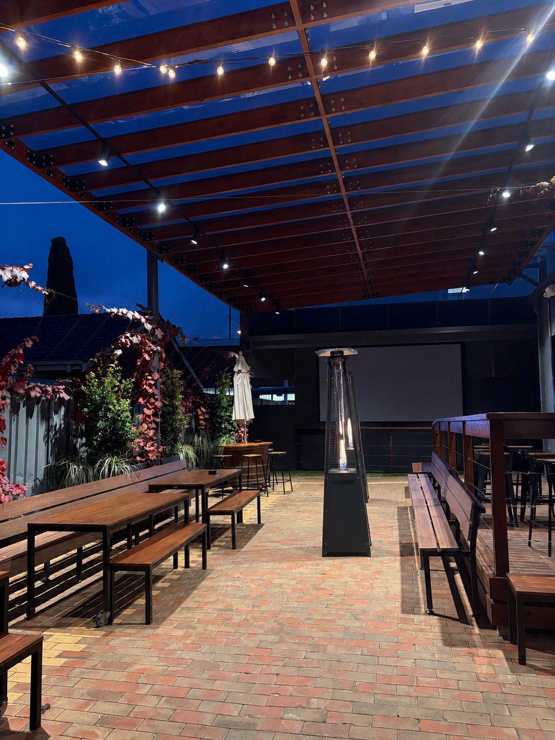 An outdoor dining patio at night with wooden benches and tables, string lights overhead, potted plants along the fence, a large patio heater, and a brick floor, possibly at a restaurant or bar.