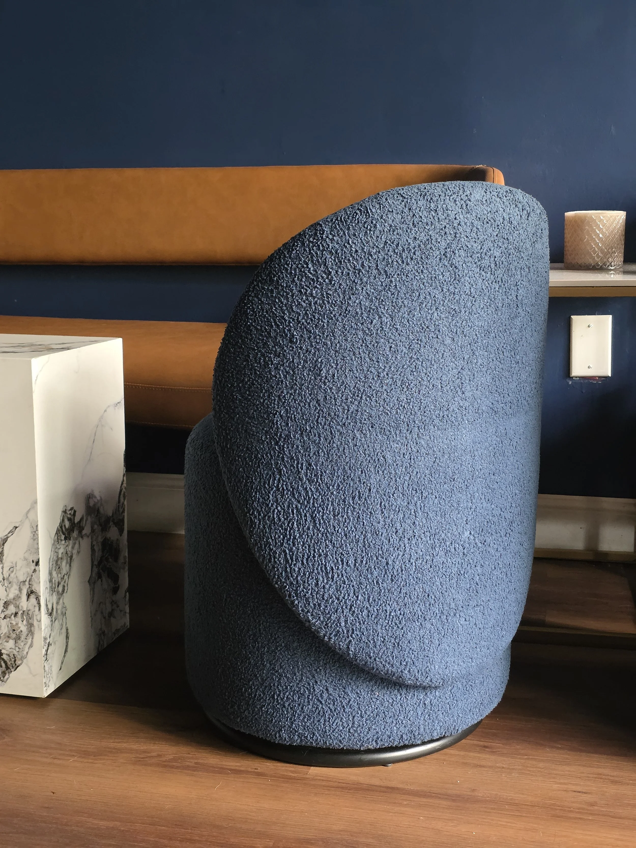 A modern living room corner featuring a rounded blue textured chair, a white marble side table, a leather bench, and a dark blue wall with a small beige candle on a shelf.