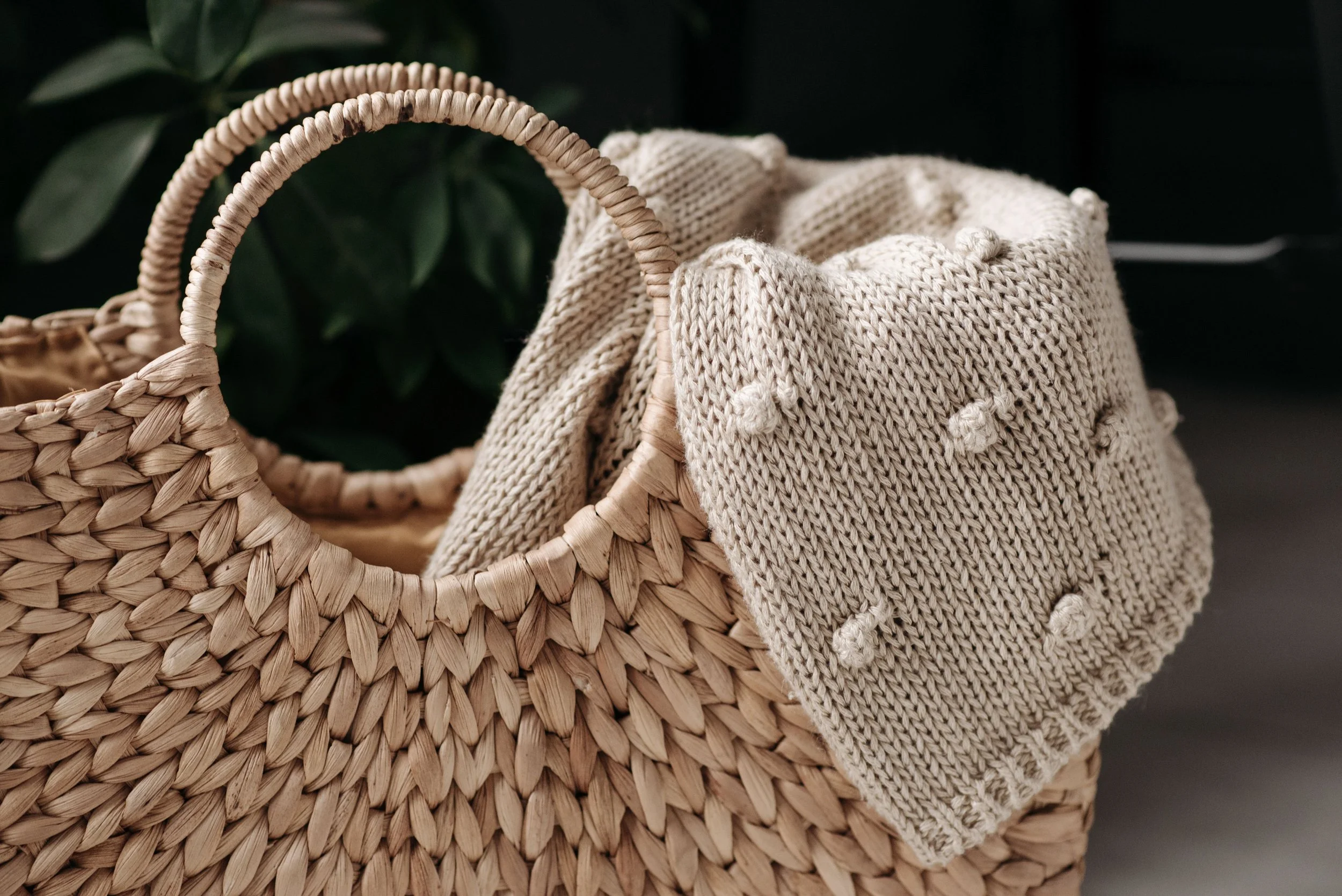 A woven basket with a handle, containing a beige knit blanket with small pom-poms, and a background of green leaves.