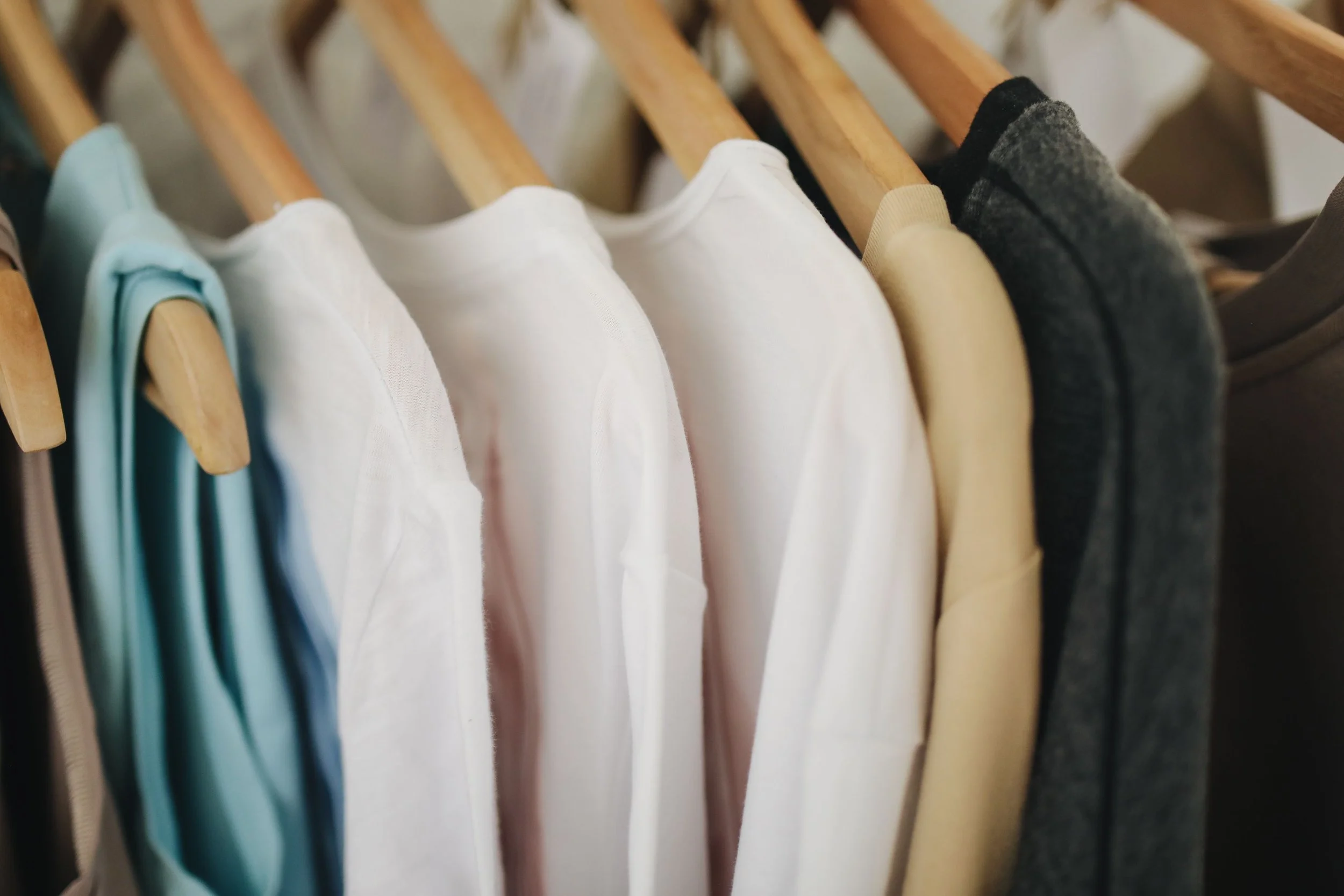 Clothes hanging on wooden hangers, including white, beige, and dark-colored shirts or sweatshirts, in a wardrobe or closet.