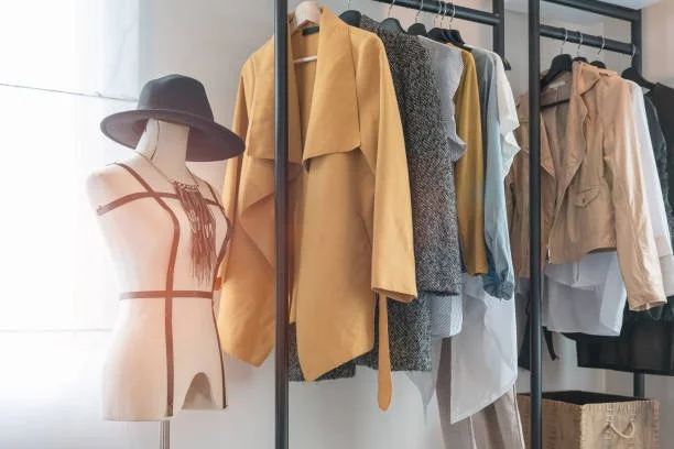 Clothing rack with assorted women's garments, including coats, shirts, and dresses, with a mannequin wearing a hat in front of a window.