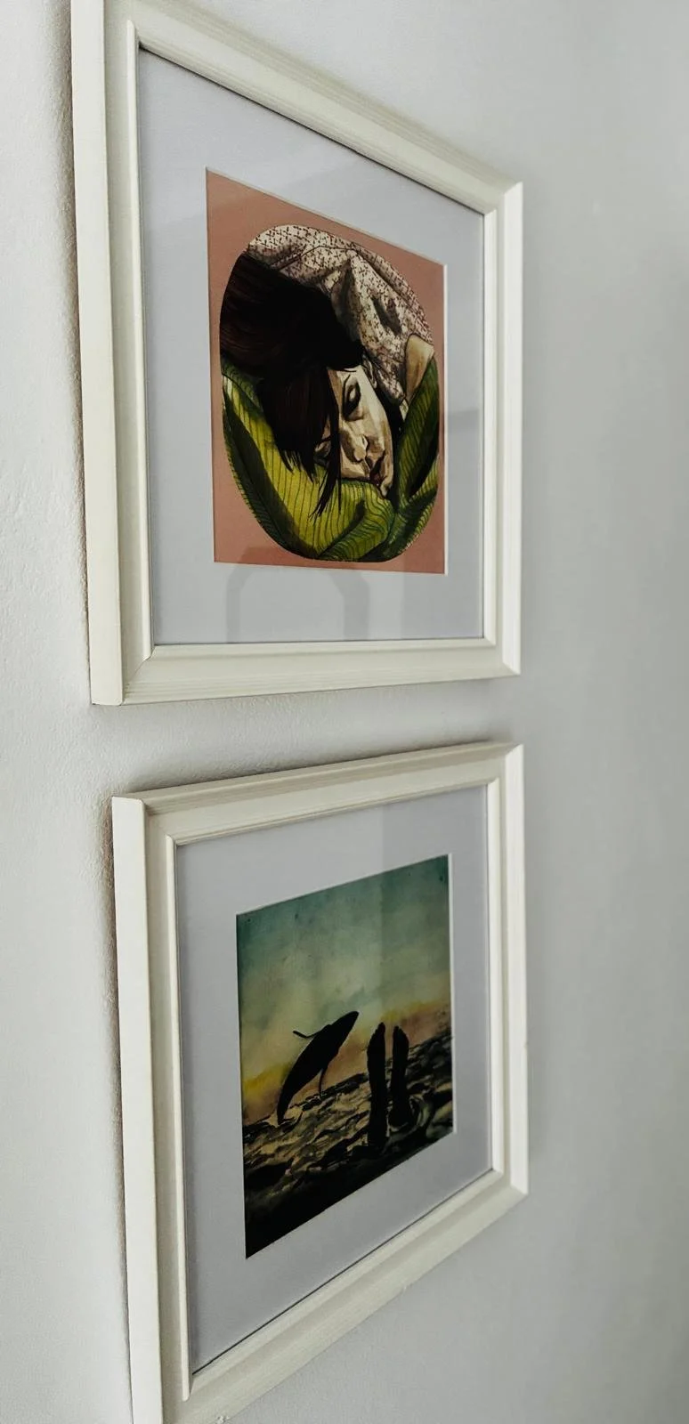 Two framed art prints hanging on a white wall. The top print depicts a woman with dark hair resting with her eyes closed, enveloped in an abstract green and pink background. The bottom print shows a landscape with dolphins jumping out of the water at sunset.