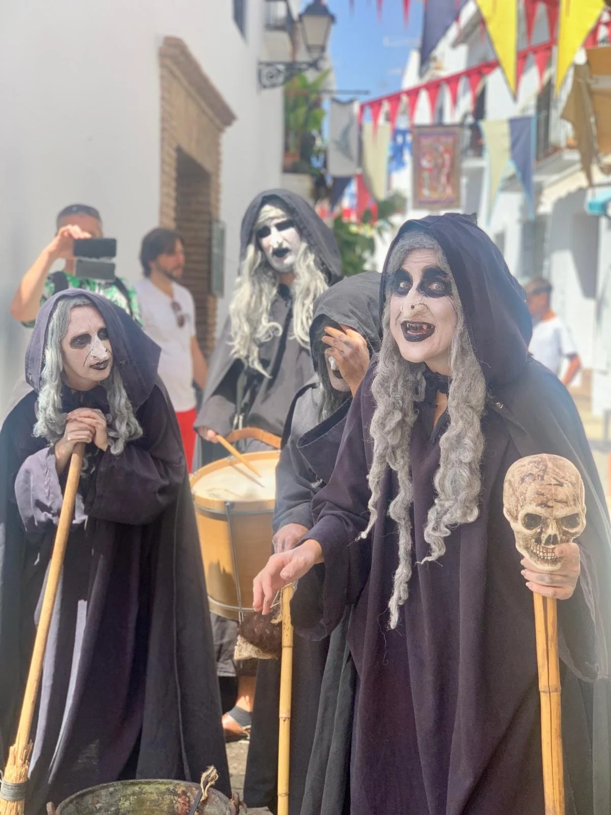 People dressed as witches or skeletons with masks, dark clothing, and staffs walking in a street decorated with colorful bunting, likely during a Halloween celebration.