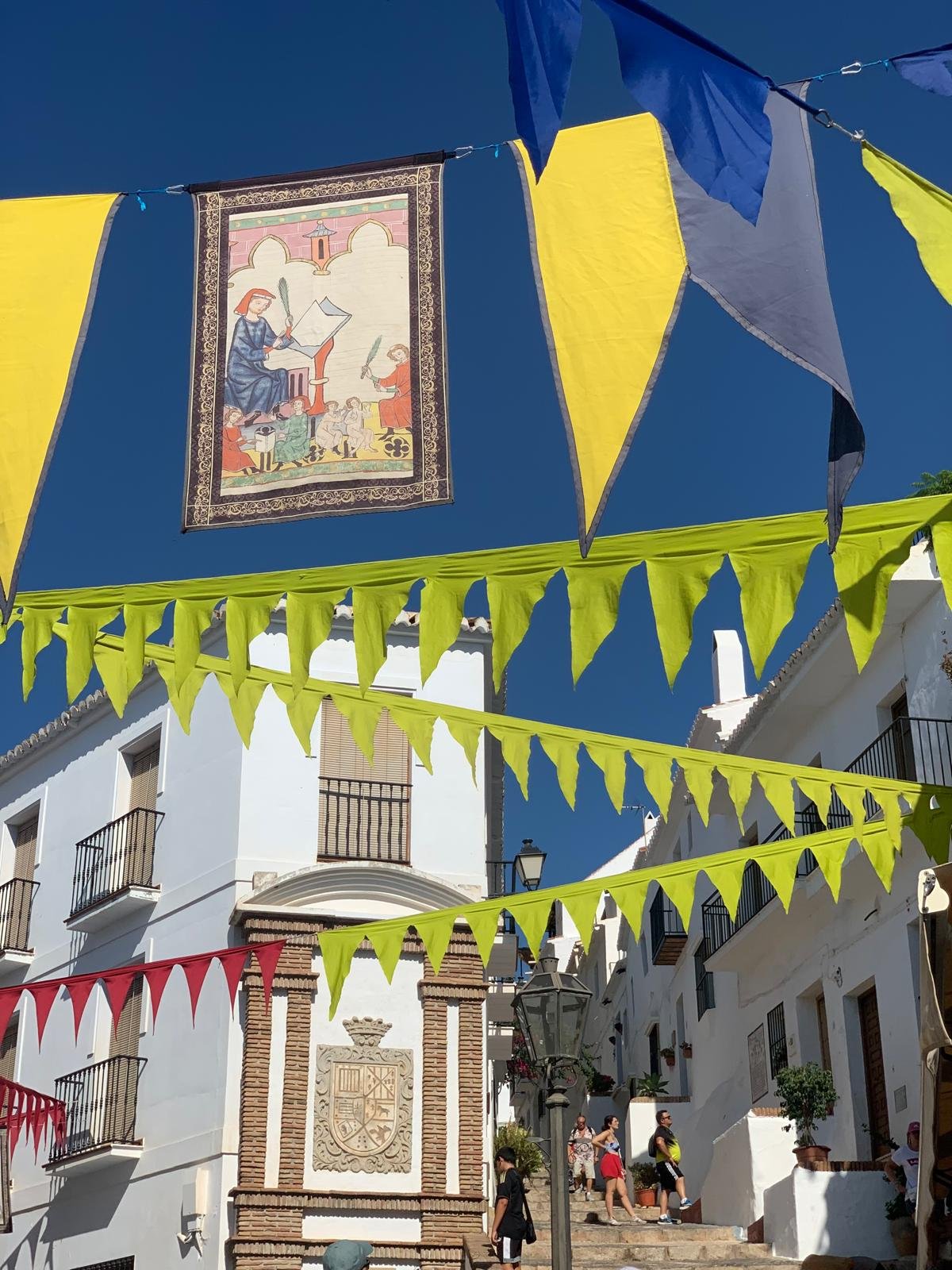White buildings with balconies and a decorative crest, string banners of yellow, blue, and red flags hanging in a sunny outdoor street scene, with people walking up and down stairs.