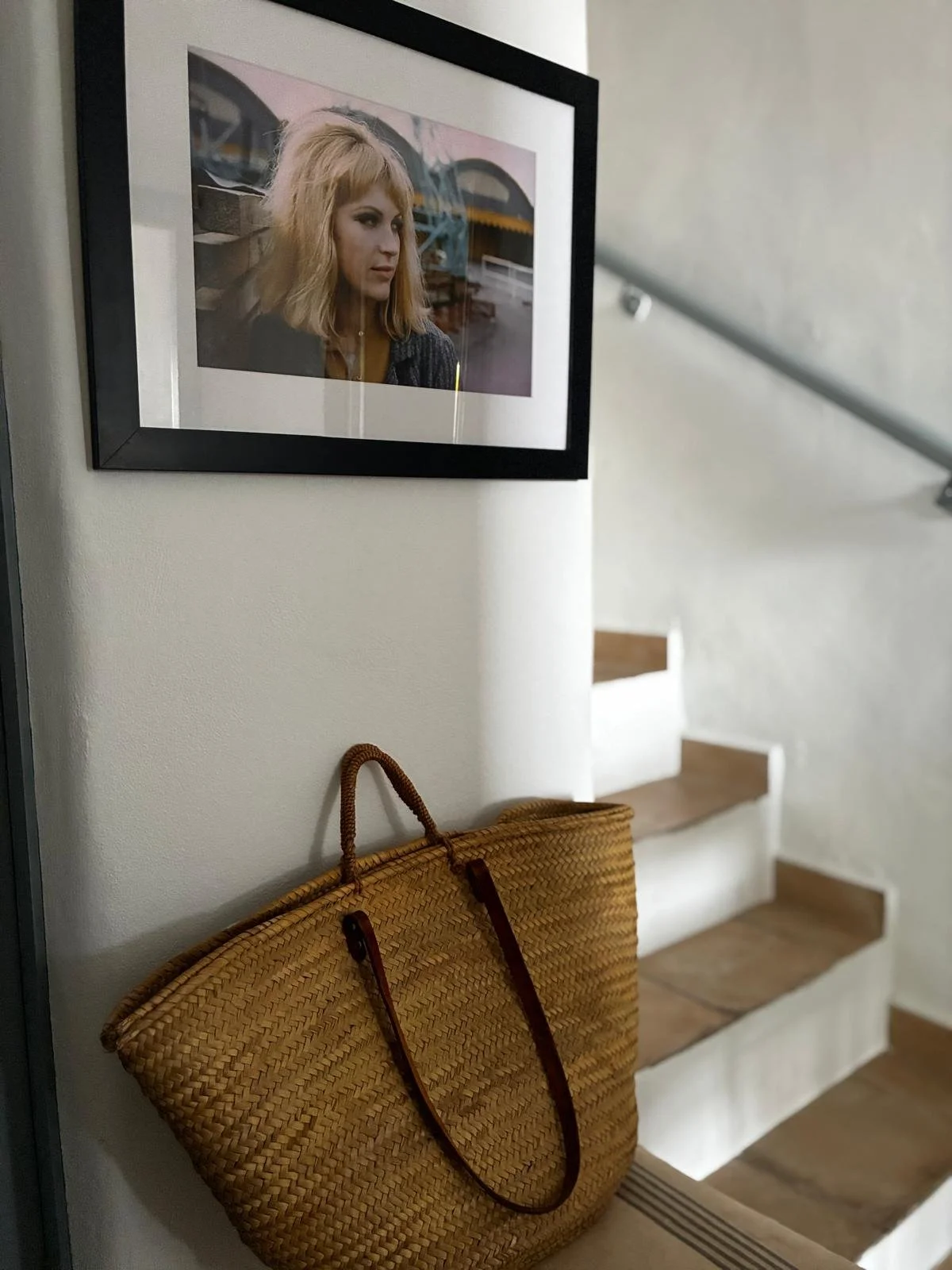 A framed photograph of a woman with blonde hair hangs on a white wall above a small wooden staircase. A large woven tote bag with leather straps leans against the wall below the photograph.