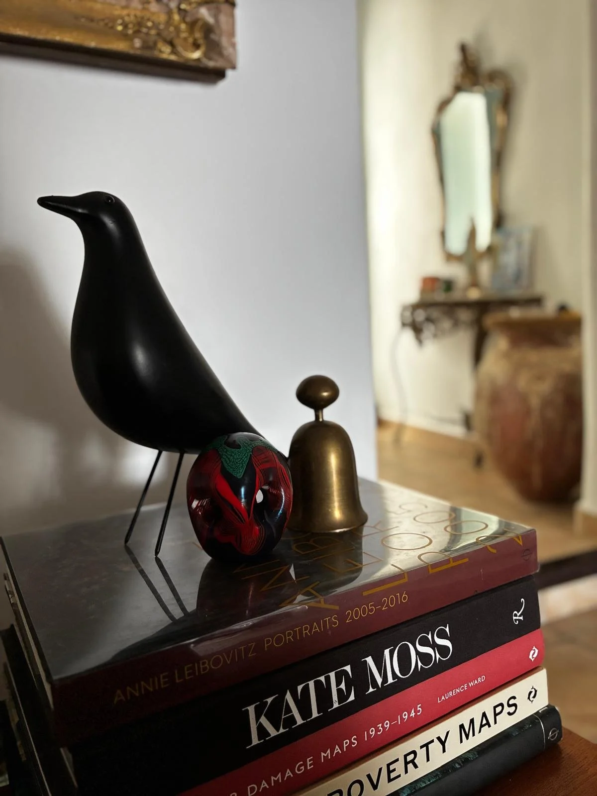 Decorative black bird sculpture, red and black glass art piece, small brass bell on top of a stack of art and history books in a cozy, ornate room.