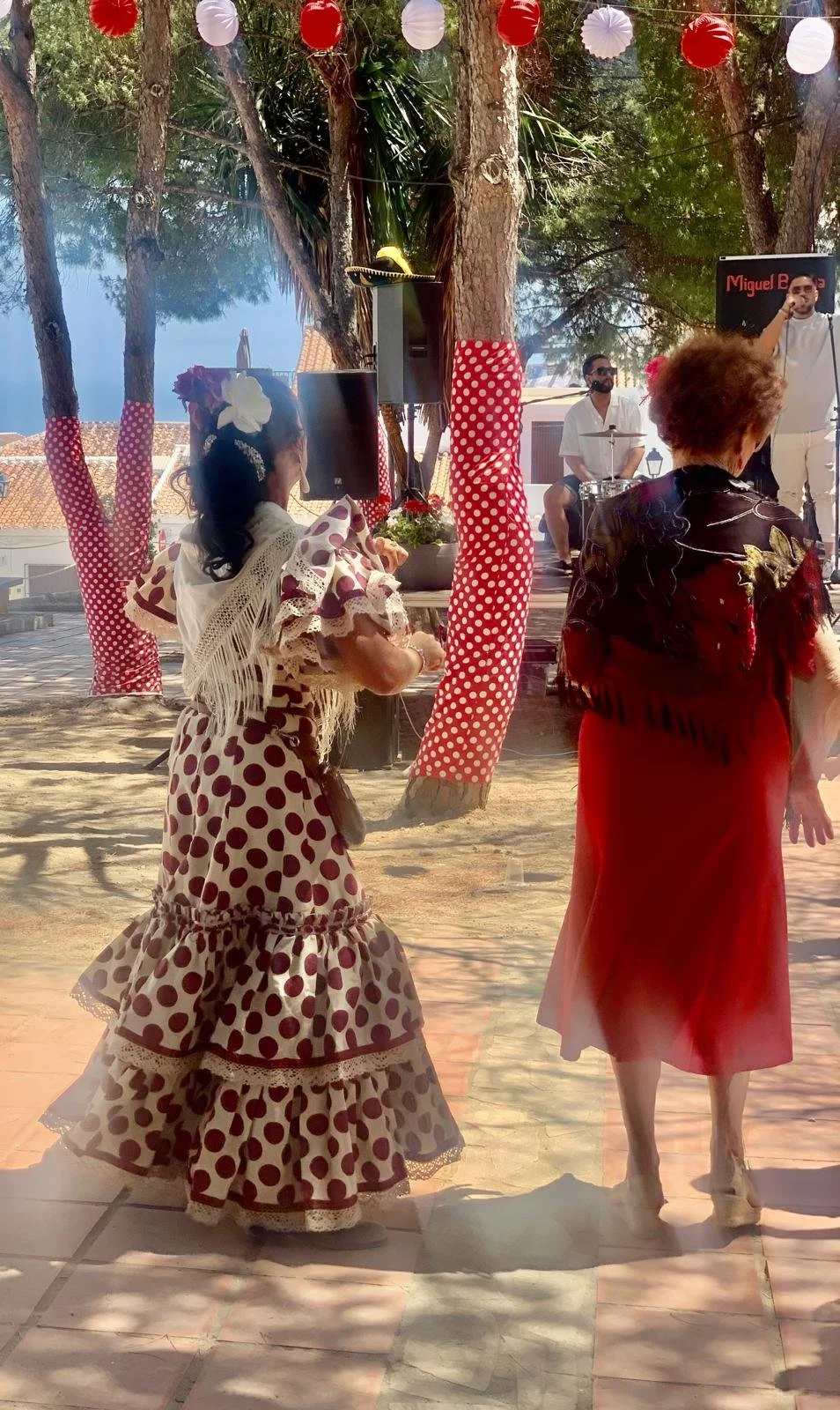Women in flamenco dresses dance outdoors under trees decorated with red and white paper lanterns while a band performs in the background.