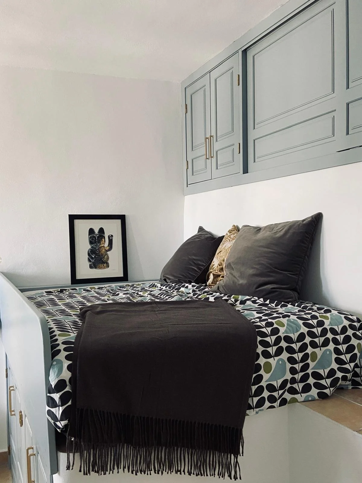 A bedroom with a bed, pillows, and a framed picture of a black cat on the wall. There are gray cabinets above the bed.