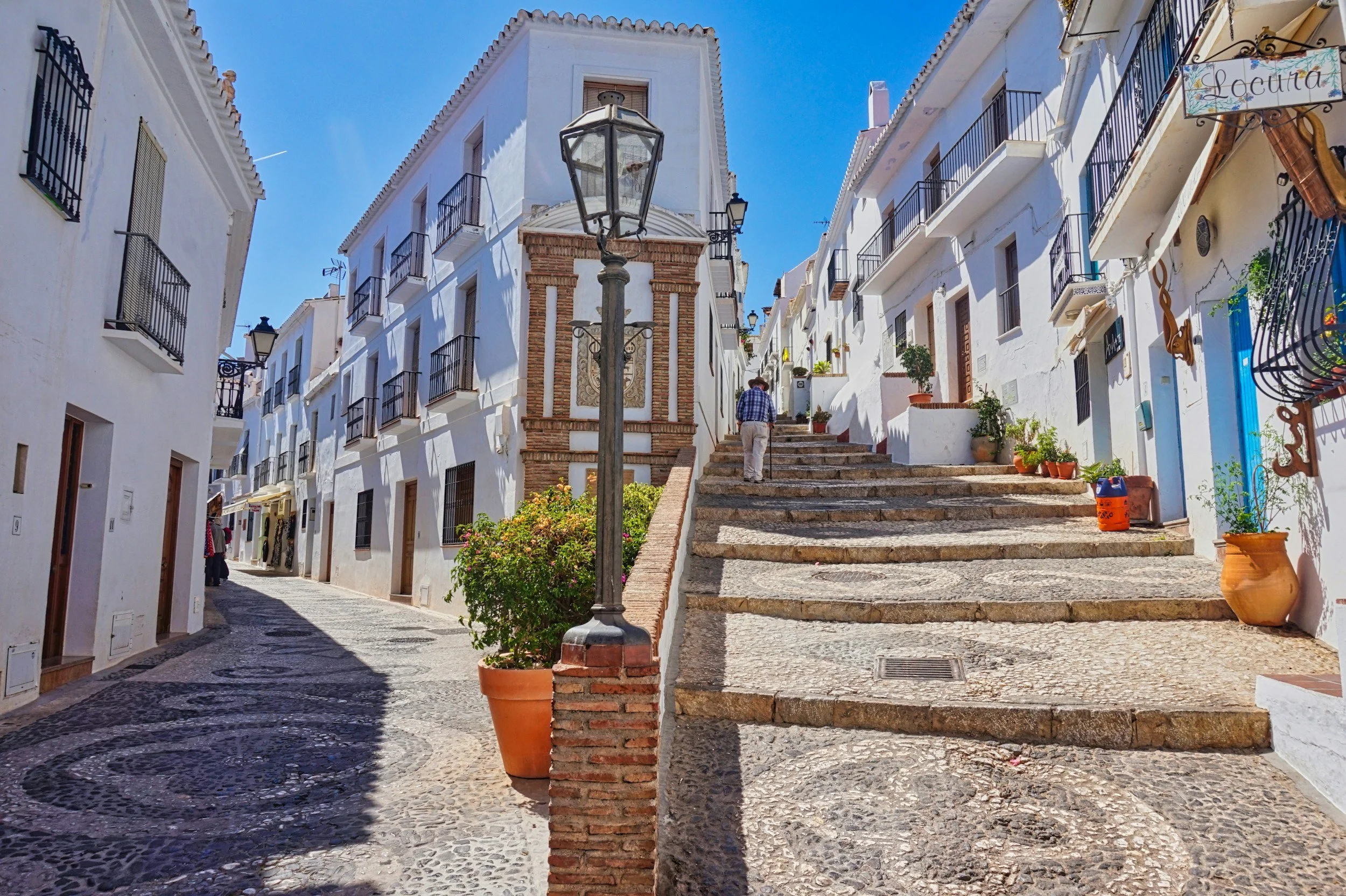 A narrow street in a Frigiliana with white buildings, potted plants, steps leading into the old town under a bright blue sky.