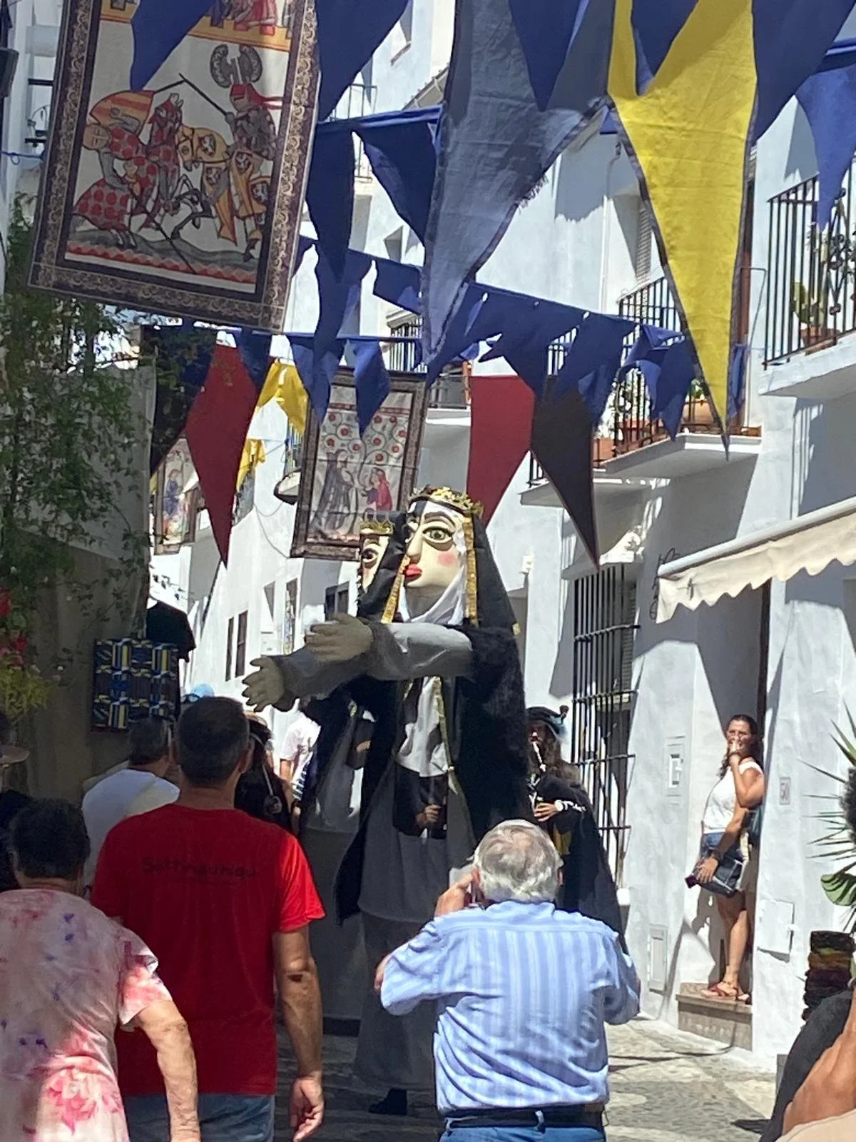 A street parade with a large puppet of a woman with long black hair, wearing traditional clothing and a crown. The puppet is surrounded by colorful flags and hanging tapestry banners. People are watching the parade in a narrow street with white buildings and balconies.