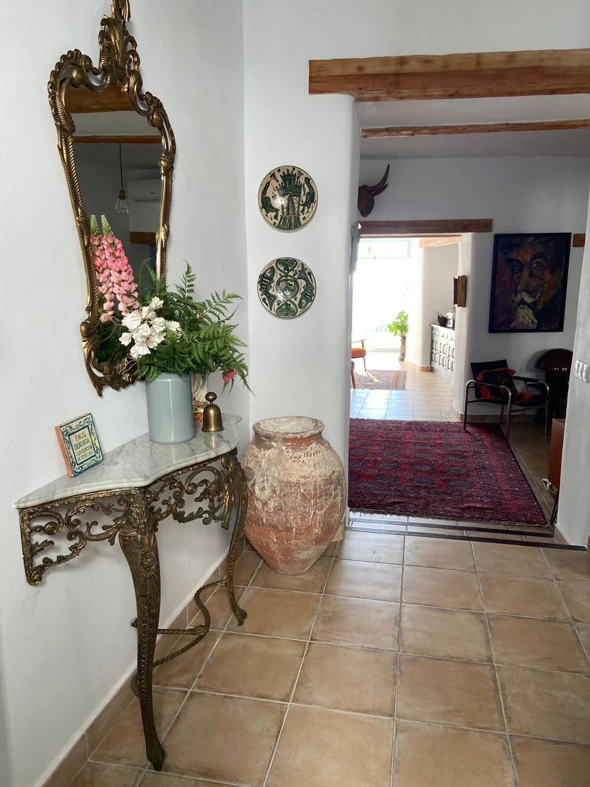 Decorative entryway with gold-framed mirror, marble-topped table, flower arrangement, large clay jar, oryx skull wall decor, and a view into a warmly lit living space with a red rug and artwork.