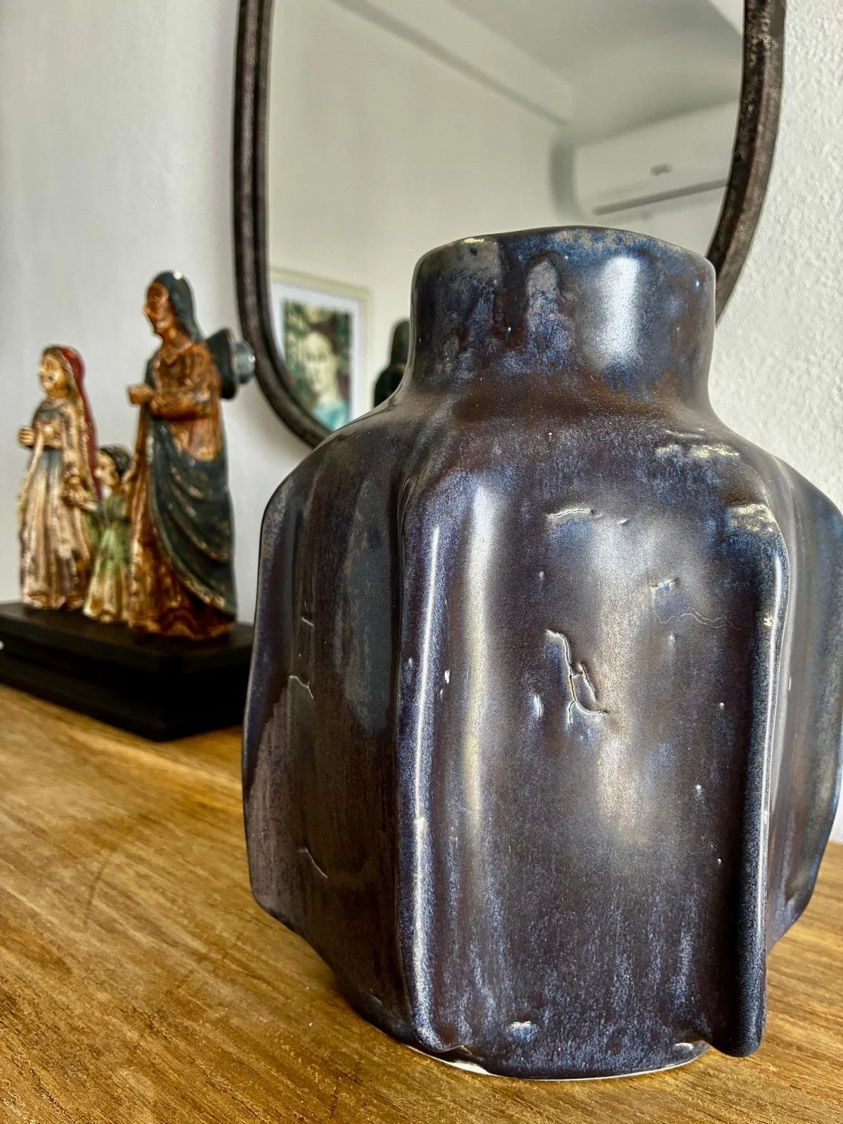 Close-up of a dark ceramic sculpture with an abstract human form on a wooden surface, with decorative statues and a mirror in the background.