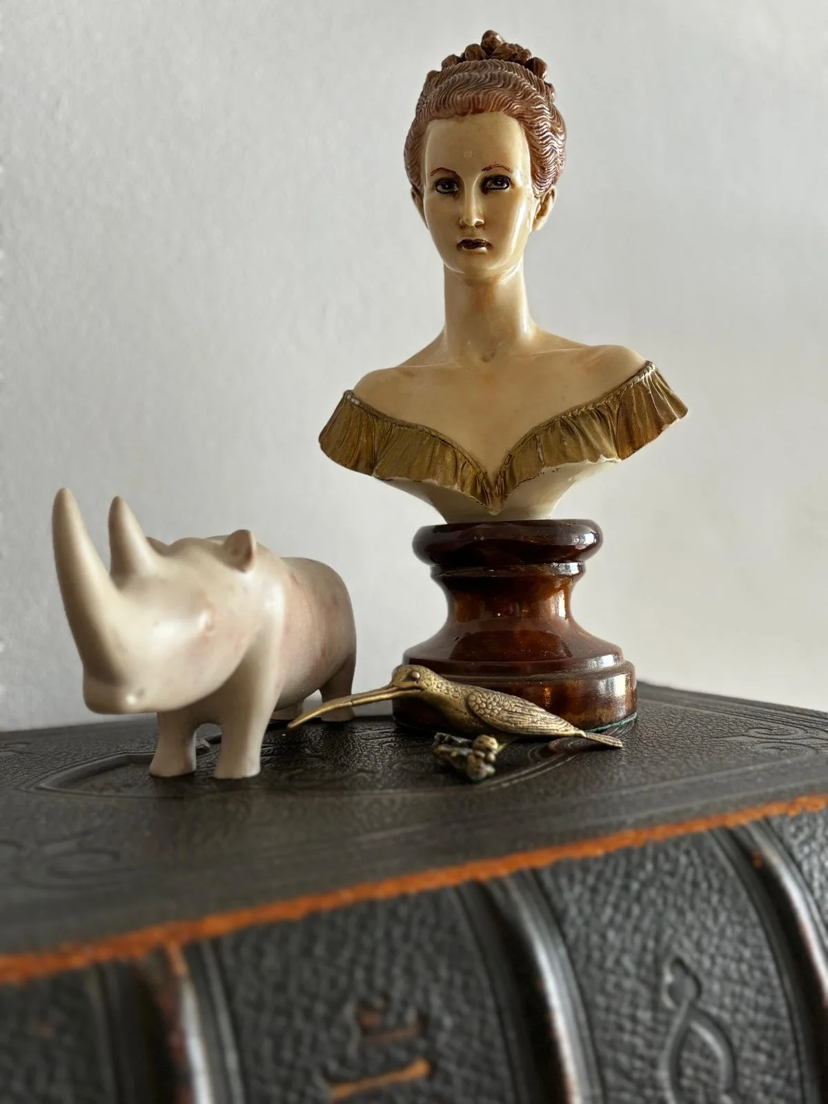 Decorative collection on a black surface: a beige rhinoceros figurine, a vintage-style bust of a woman with reddish hair in an updo, a small bronze-colored bird figurine, and a tiny frog figurine.