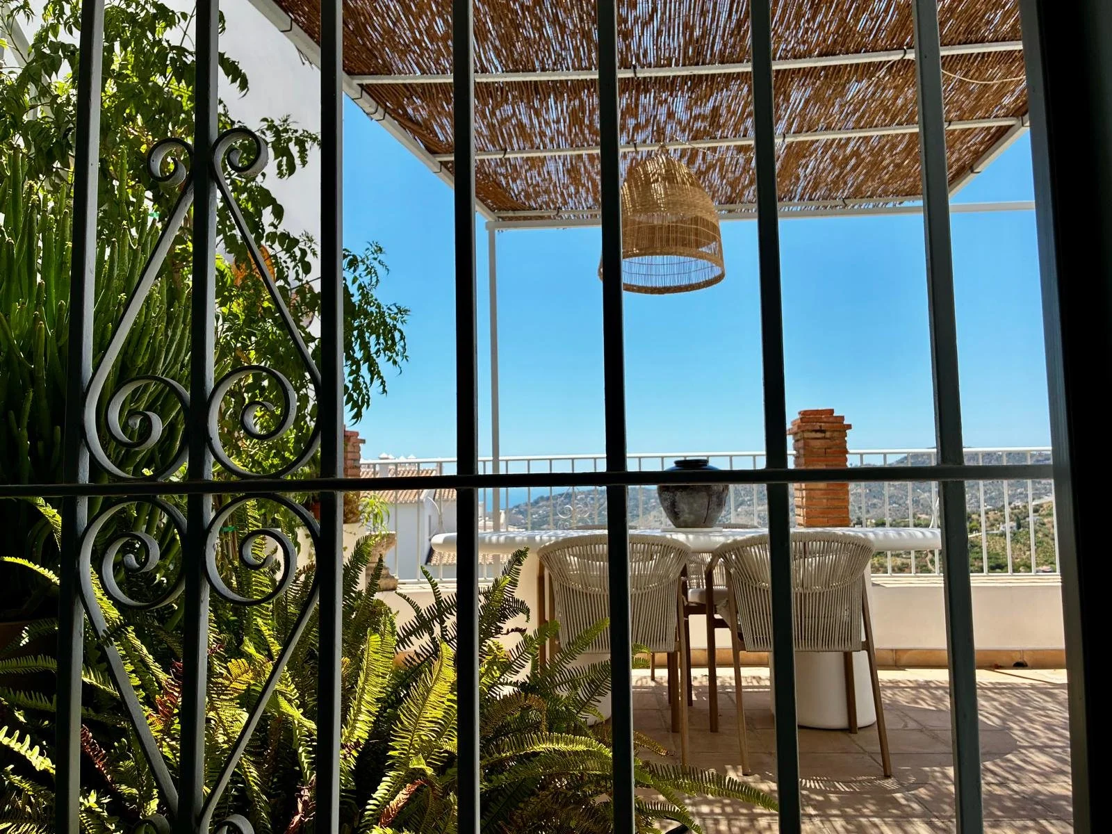 View of a balcony with outdoor furniture, greenery, a thatched roof, and a blue sky, seen through black wrought iron bars.
