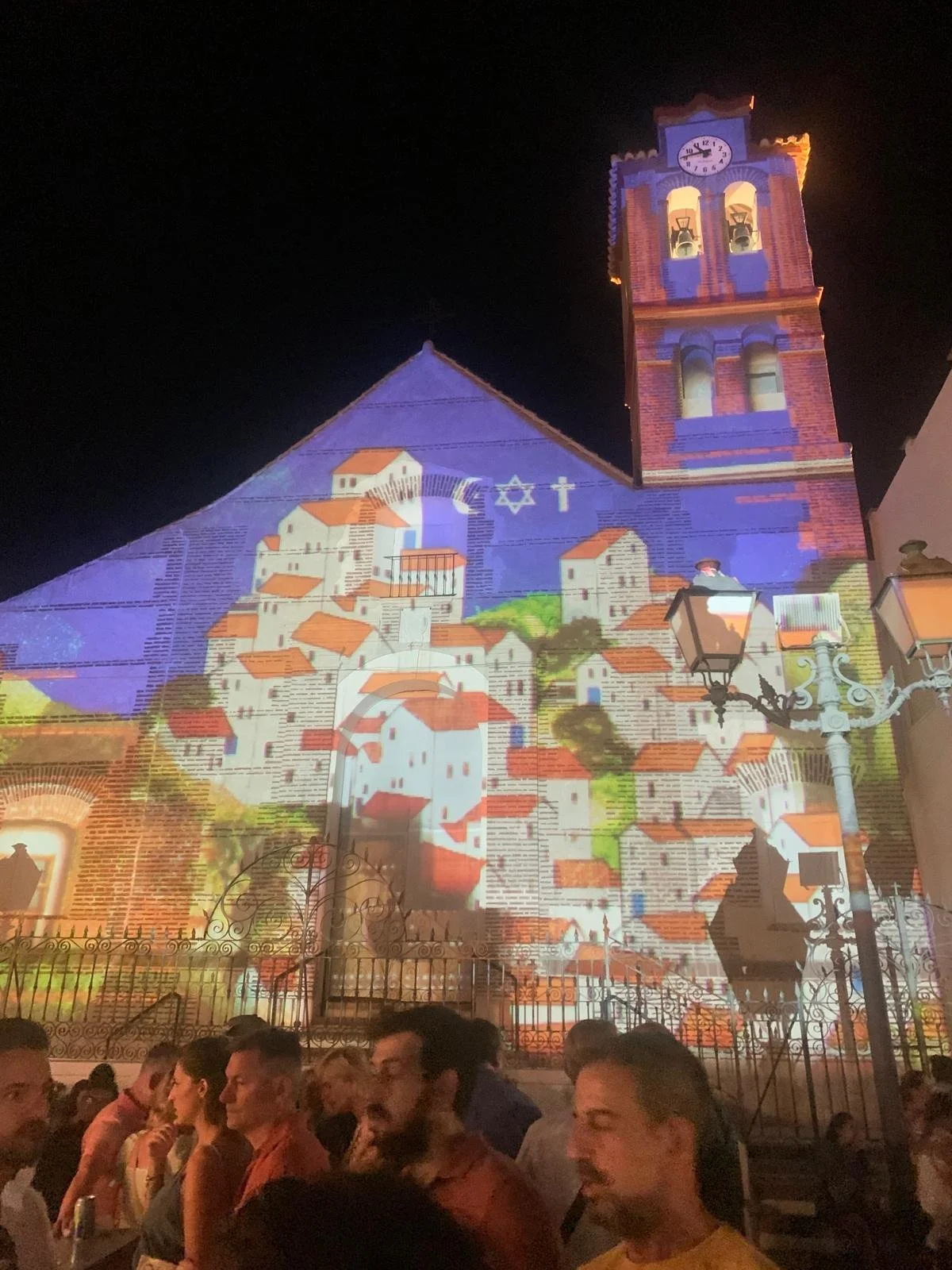 Nighttime projection on St Antonio church in Frigiliana showing a scene with houses, religious symbols, and colourful artwork, with a crowd of people in the foreground.