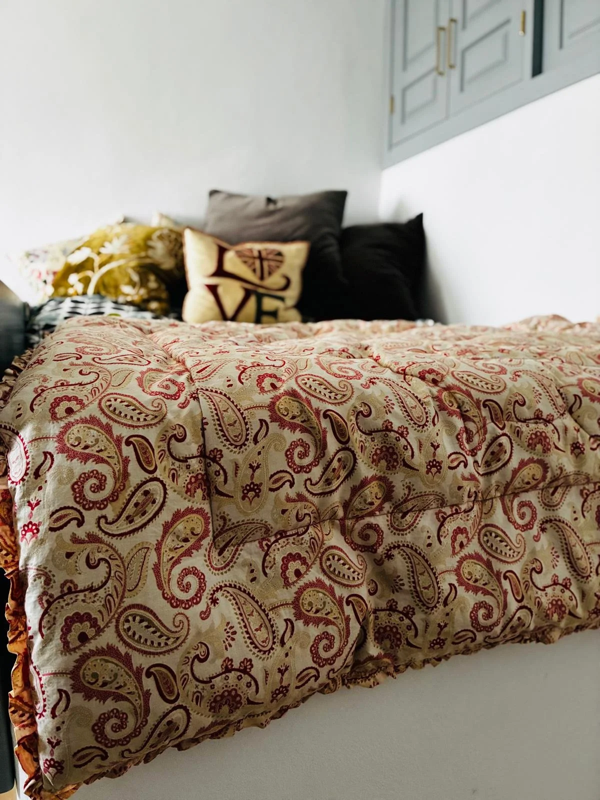 A bed with a paisley-patterned quilt, several pillows including one with the word "LOVE" and a heart, and a white wall in the background.