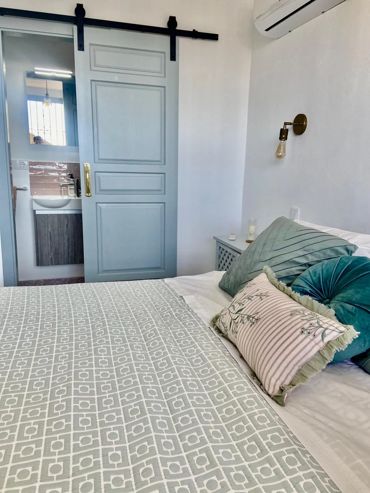 Bedroom with neatly made bed, decorative pillows, bedside table with personal items, wall-mounted light fixture, air conditioning unit, and sliding door leading to adjacent bathroom with sink and mirror.