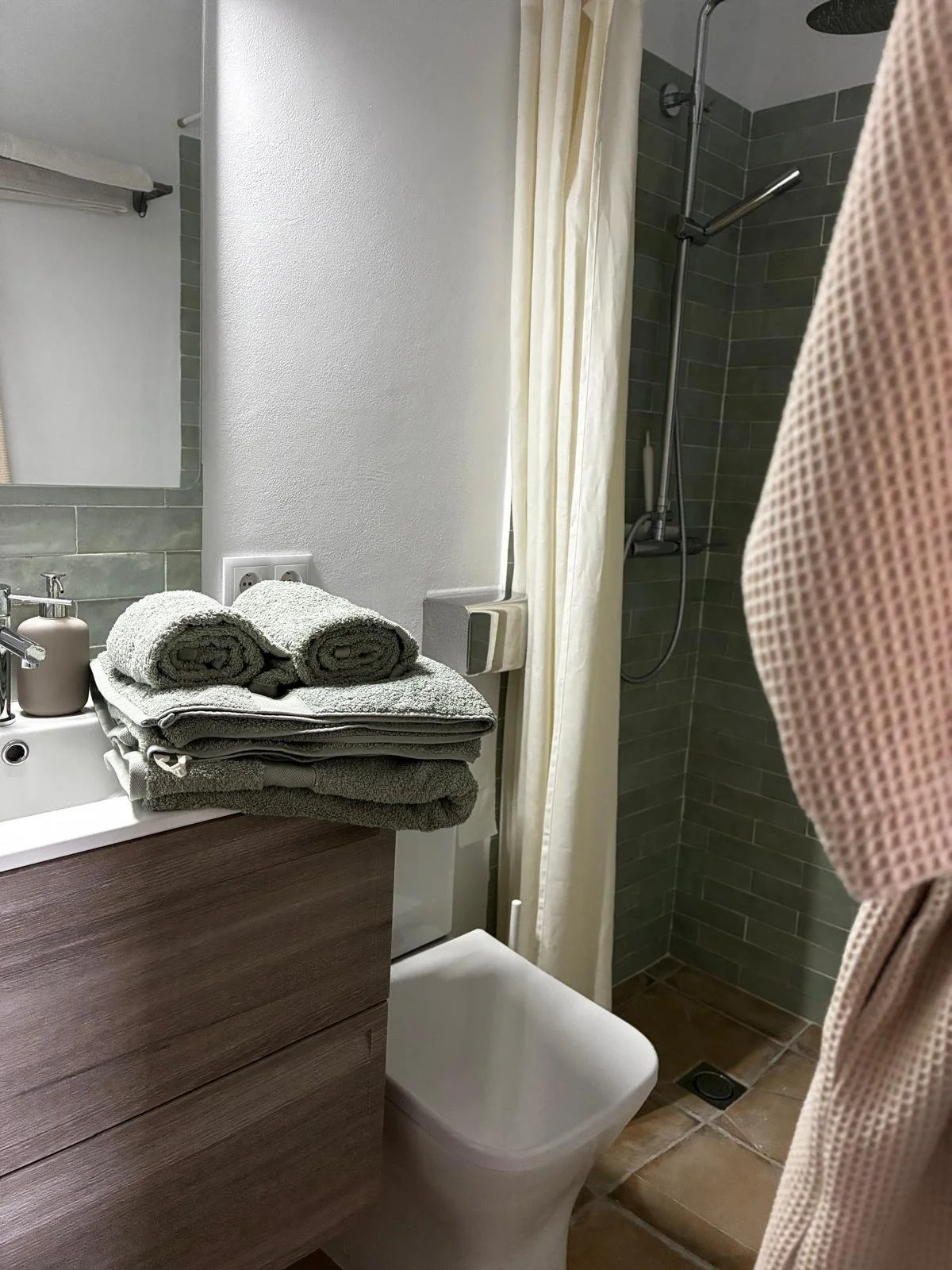 A bathroom with a sink, mirror, and towels on the counter. A shower with a curtain and tiled walls is visible in the background.