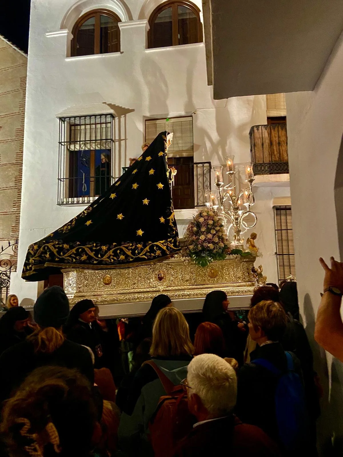 A religious procession at night with a float carrying a statue of the Virgin Mary, covered with a black cloth adorned with gold stars. The float is decorated with flowers and candelabras, and is carried by people among a crowd gathered around.