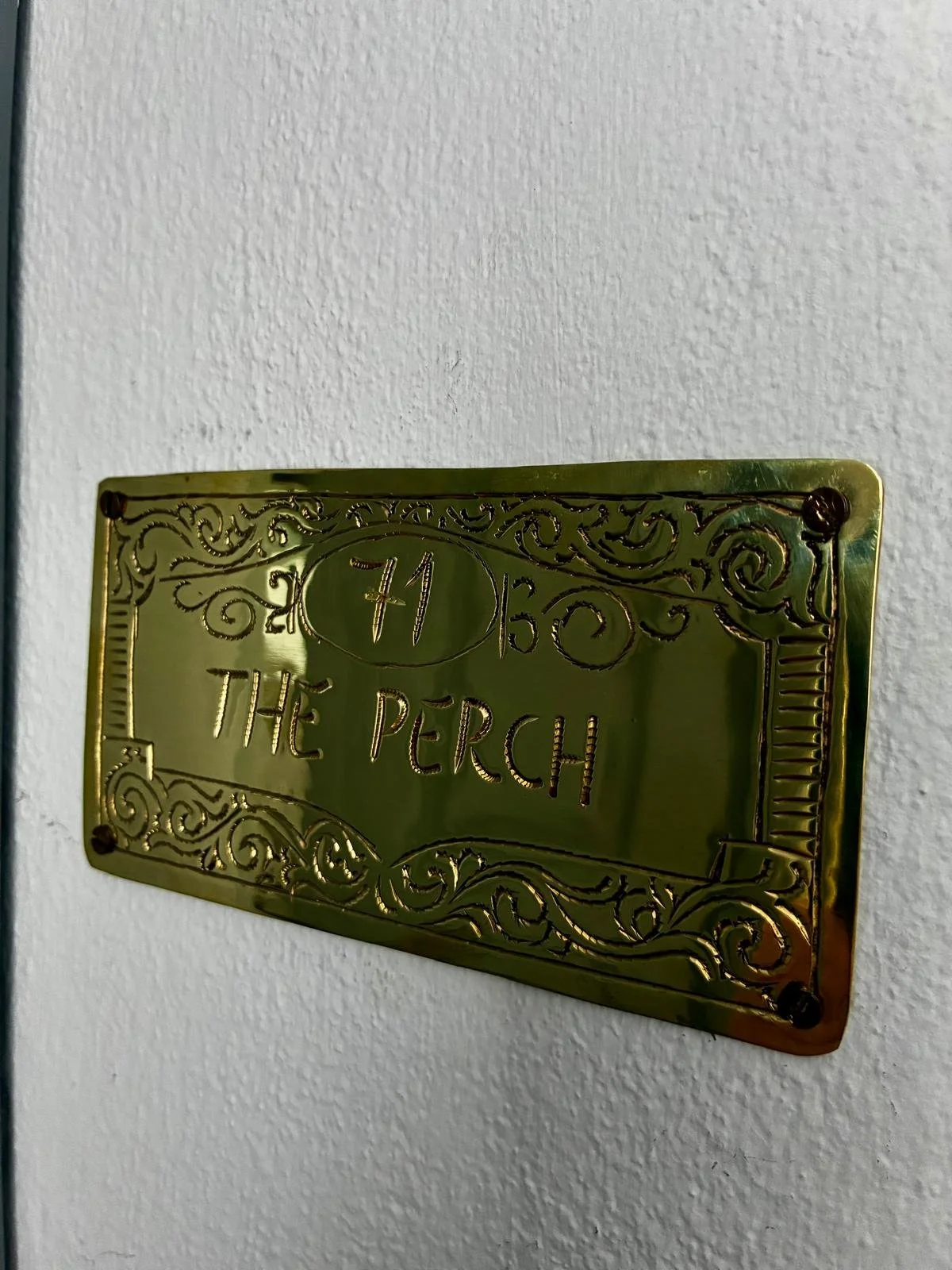 A decorative brass plaque on a white wall, engraved with the words "The Perch" and the number "41" inside an oval, with ornate scrollwork designs surrounding the text.