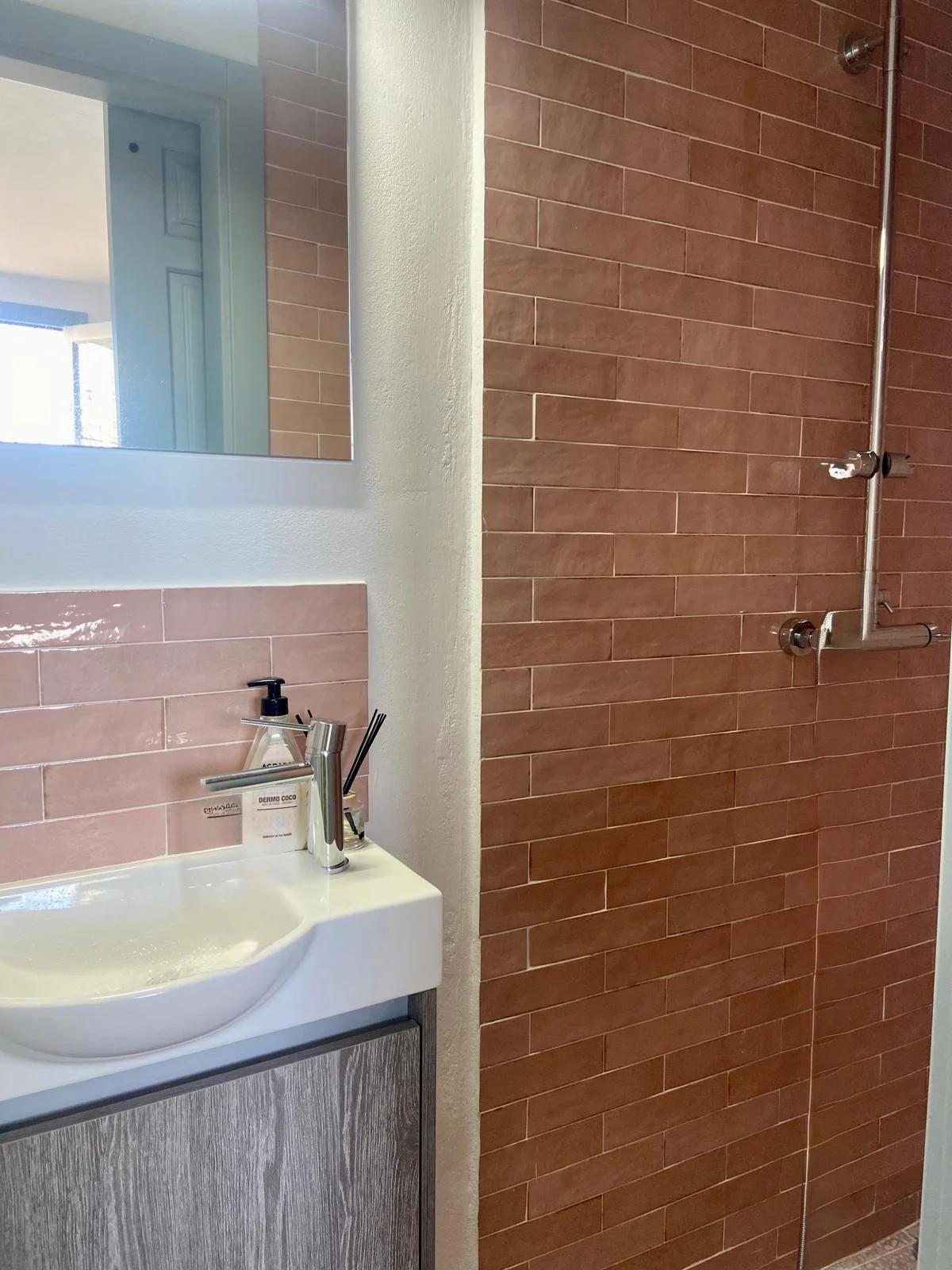 Bathroom with a rectangular mirror above a sink, pink tiles behind the sink, and a brown-tiled shower area with a metal shower pipe and controls.