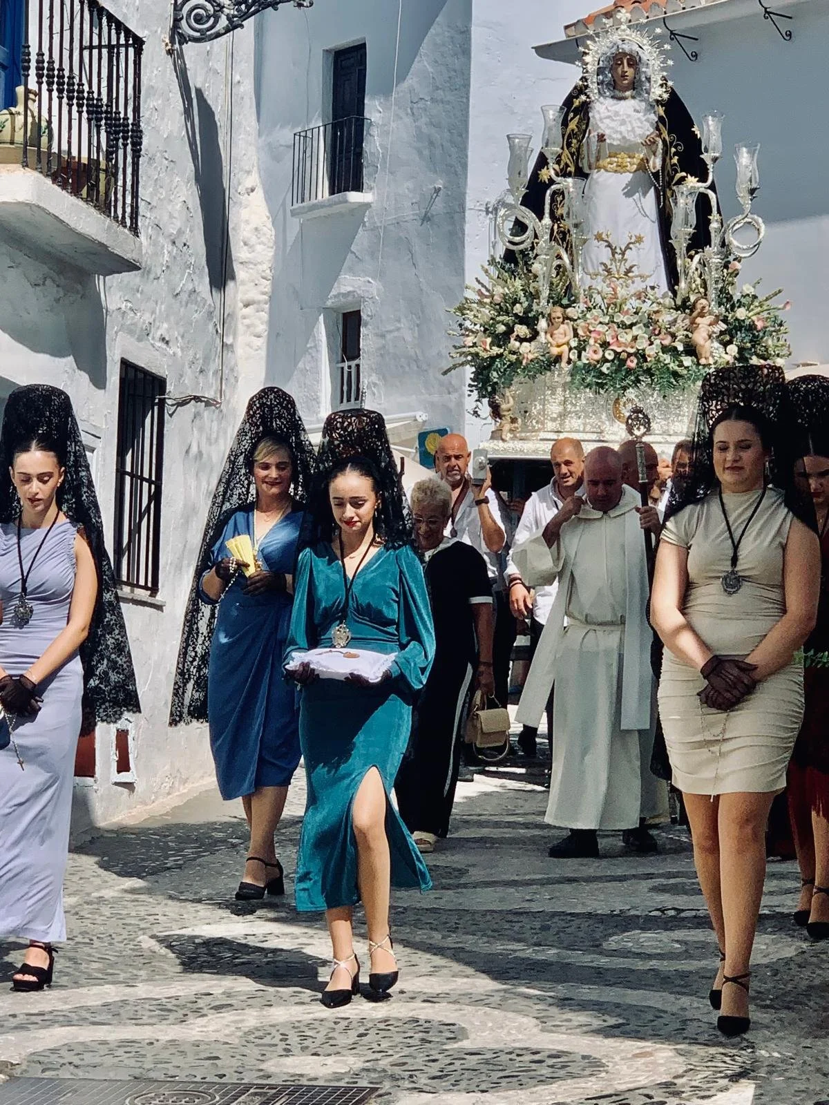 People participating in a religious procession carrying a religious statue decorated with flowers through a narrow street with whitewashed buildings