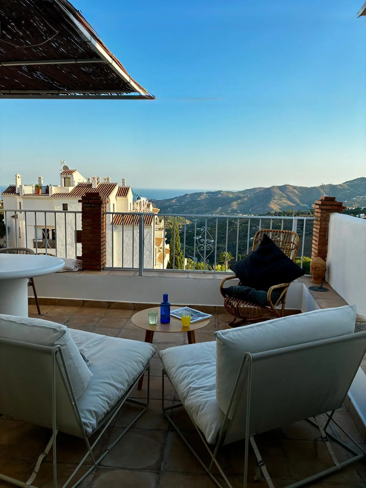 A balcony with a view of mountains and white buildings with red tile roofs. The balcony has white cushioned chairs, a small round wooden table with drinks and a magazine, and a rattan chair with a black cushion. There is a terracotta sculpture of a head on a ledge and a decorative white railing.