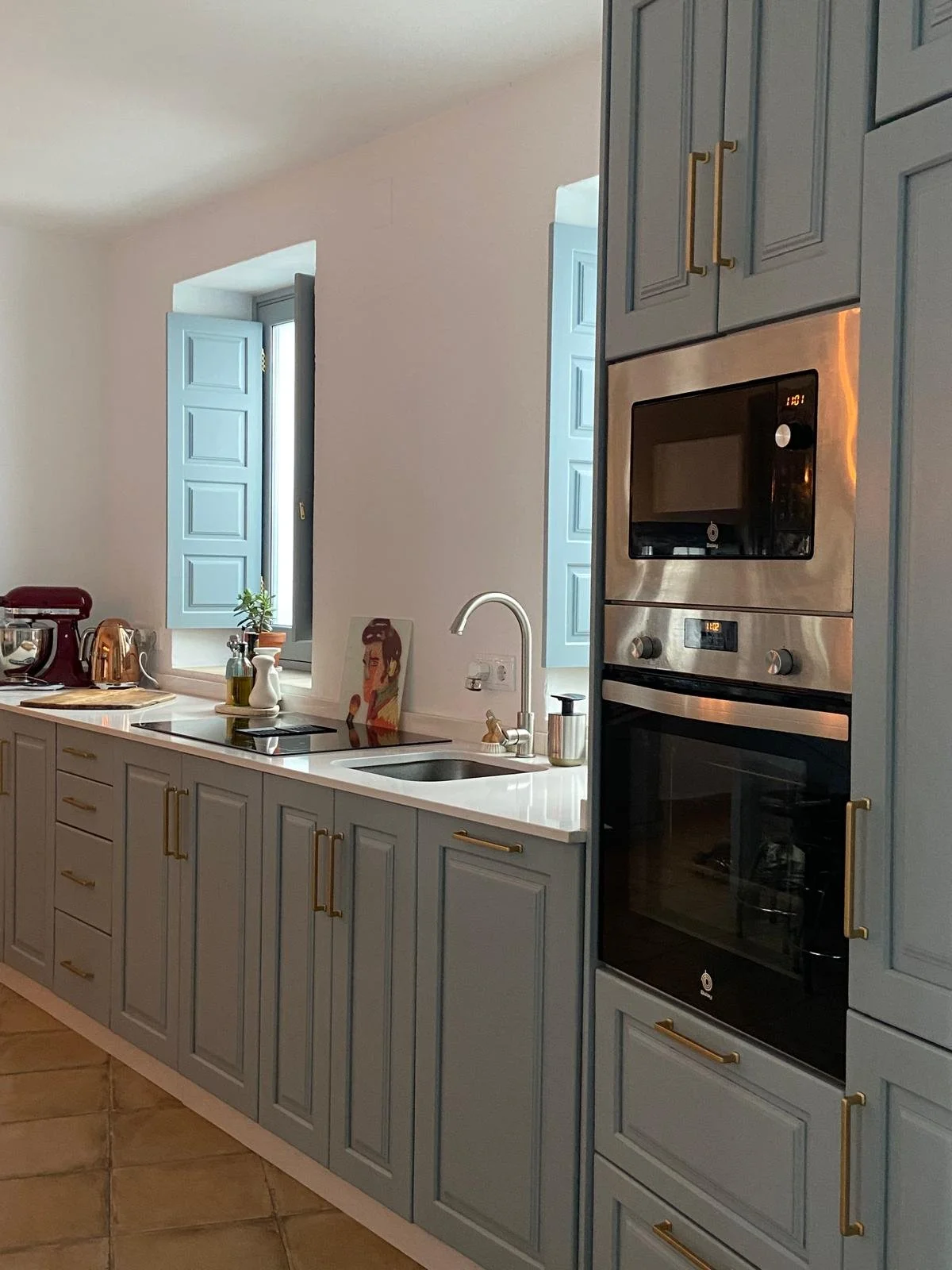 Modern kitchen with light blue cabinets, gold hardware, a white countertop, a sink, a window with light blue shutters, a small potted plant, a painting, and built-in stainless steel oven and microwave.
