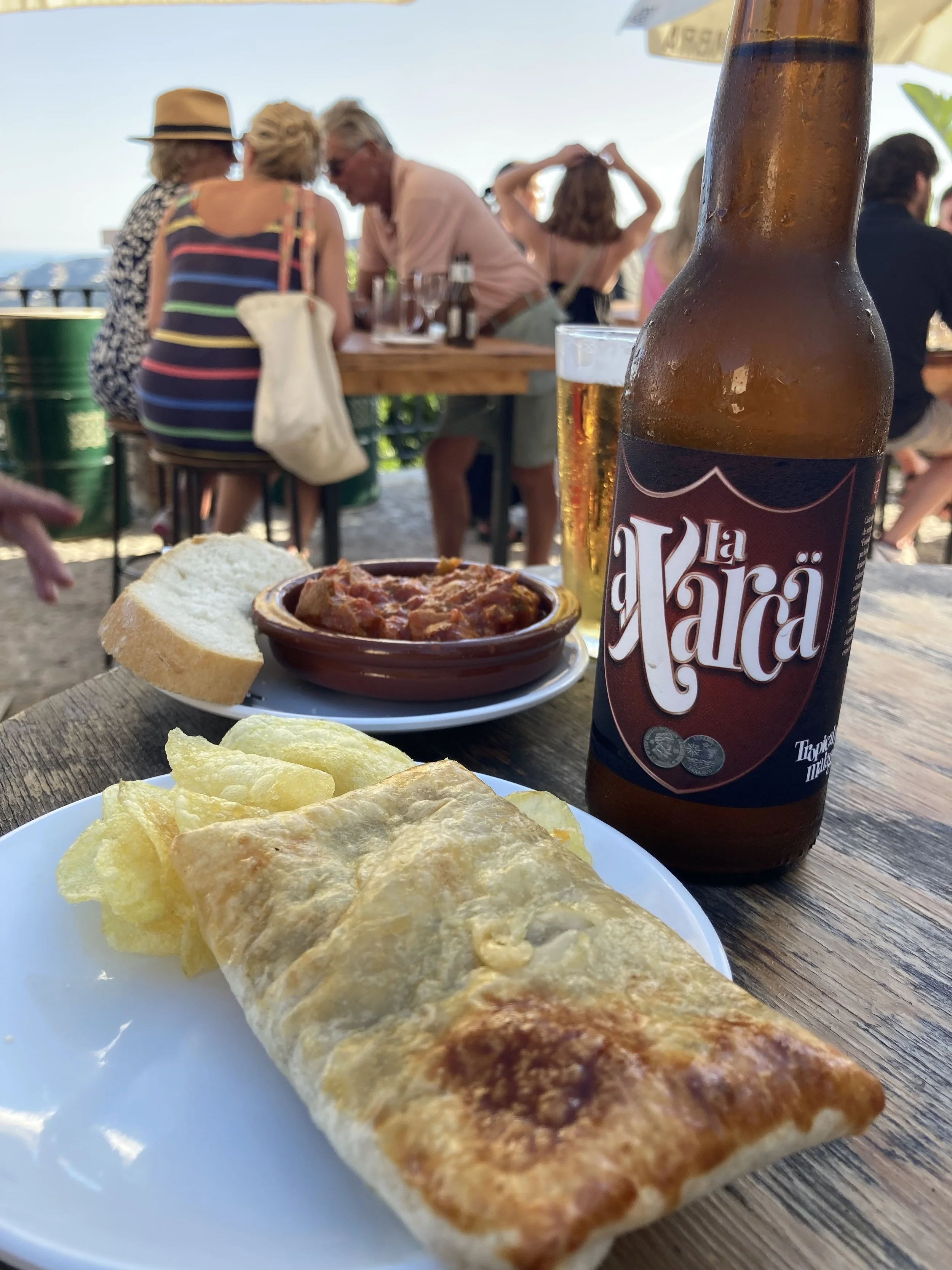 A picture of tapas and a bottle of the local Axarca craft beer in a local bar. People in the background are enjoying the view, tapas and drinks.