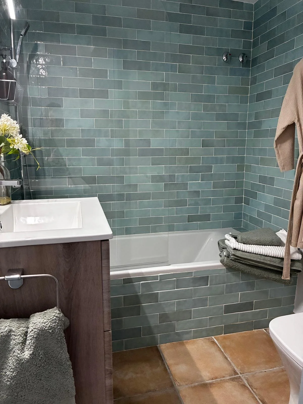 A bathroom with green subway tile walls, a white bathtub, a wooden vanity with a white sink, a towel rack with folded towels, and a beige towel hanging. There are flowers on the vanity and a brown tiled floor.